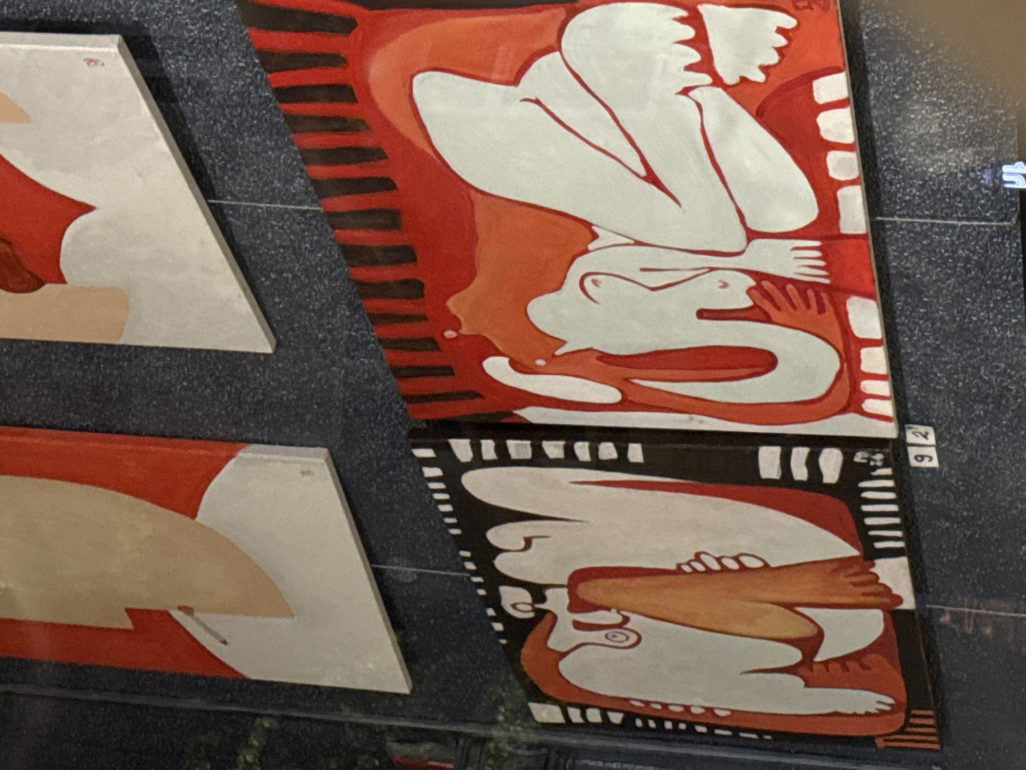 The image displays a collection of modern, abstract paintings featuring stylized human forms, set against a dark, textured background, possibly a wall or display panel. The perspective is an oblique, downward-looking shot.
Several canvases are visible. To the left, two triangular-shaped paintings show partial figures in white and beige against a red background. A small red marking, likely a signature or symbol, is faintly visible on the upper left corner of these triangular pieces.
To the right, two larger, rectangular canvases are stacked vertically. The top rectangular painting depicts white, nude, and curvaceous figures on a background of various shades of red and orange. The figures appear to be in reclining or curled poses, with exaggerated hands and feet. Black horizontal stripes are present along the left edge of this painting.
Below it, the bottom rectangular painting features similar stylized white figures, with parts in beige, set against a black background with white abstract patterns along the edges. One figure appears to be holding a beige form resembling a limb or cloth.
A small white tag with the numbers "92" is attached to the bottom right corner of the upper rectangular painting. Another tag with a faint "S" is barely visible next to the upper painting. The dark background has a speckled or grainy texture, and thin wires, possibly for hanging the art, are visible between the canvases.
The overall style of the paintings is minimalist yet expressive, using bold outlines and flat areas of color. The artwork does not provide specific location cues for Varna, Bulgaria.