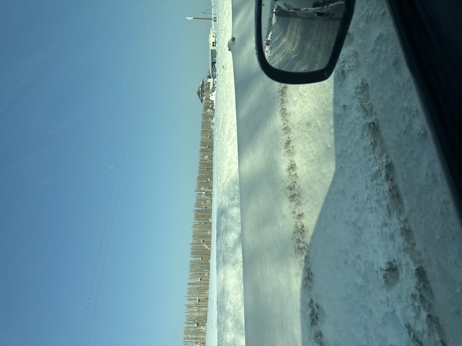 The image depicts a winter scene from a vehicle's perspective. A snow-covered road runs alongside a fence, with occasional buildings in the distance under a clear blue sky. The vehicle's side mirror is visible, reflecting the road and a hint of another car. Snow covers the ground and the road surface, with dark tracks indicating tire marks. The scene suggests a cold, possibly rural, environment.