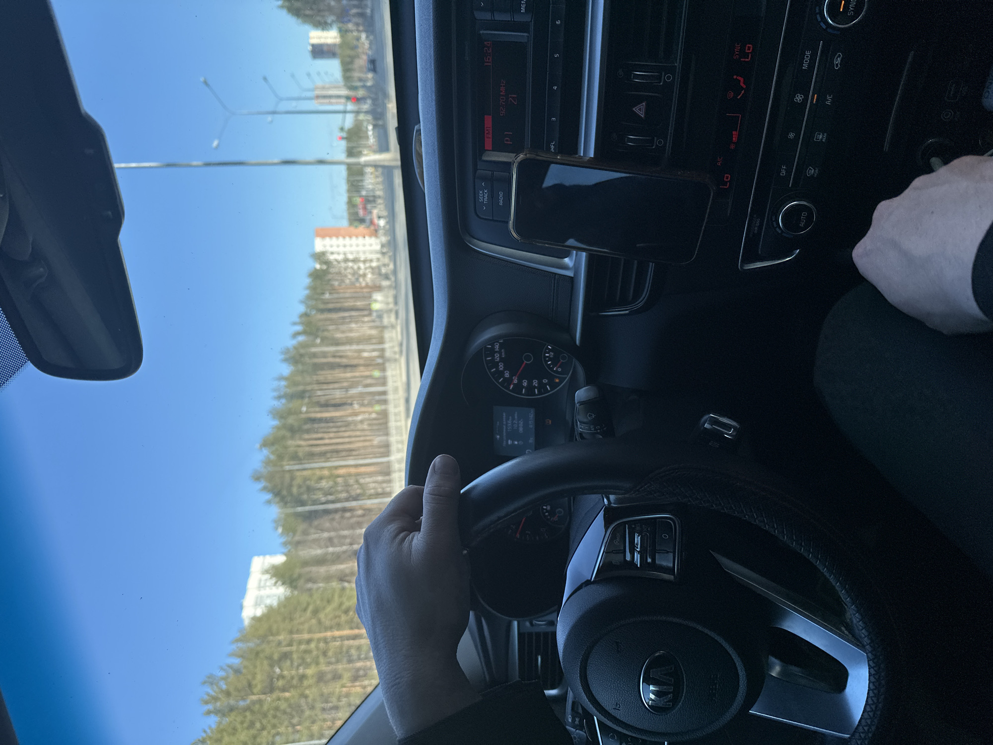 The image depicts the interior of a car, specifically the driver's perspective. A hand is visible on the steering wheel, indicating someone is driving. The dashboard shows a Kia logo on the steering wheel, a smartphone mounted near the center console, and the car radio tuned to 92.70 MHz. The display reads 16:24, and the climate control is set to "LO".
Outside the windshield, trees line the road, and in the distance are buildings, including a tall white building with orange accents. Based on the visual clues, this location appears to be in Yekaterinburg, Russia.