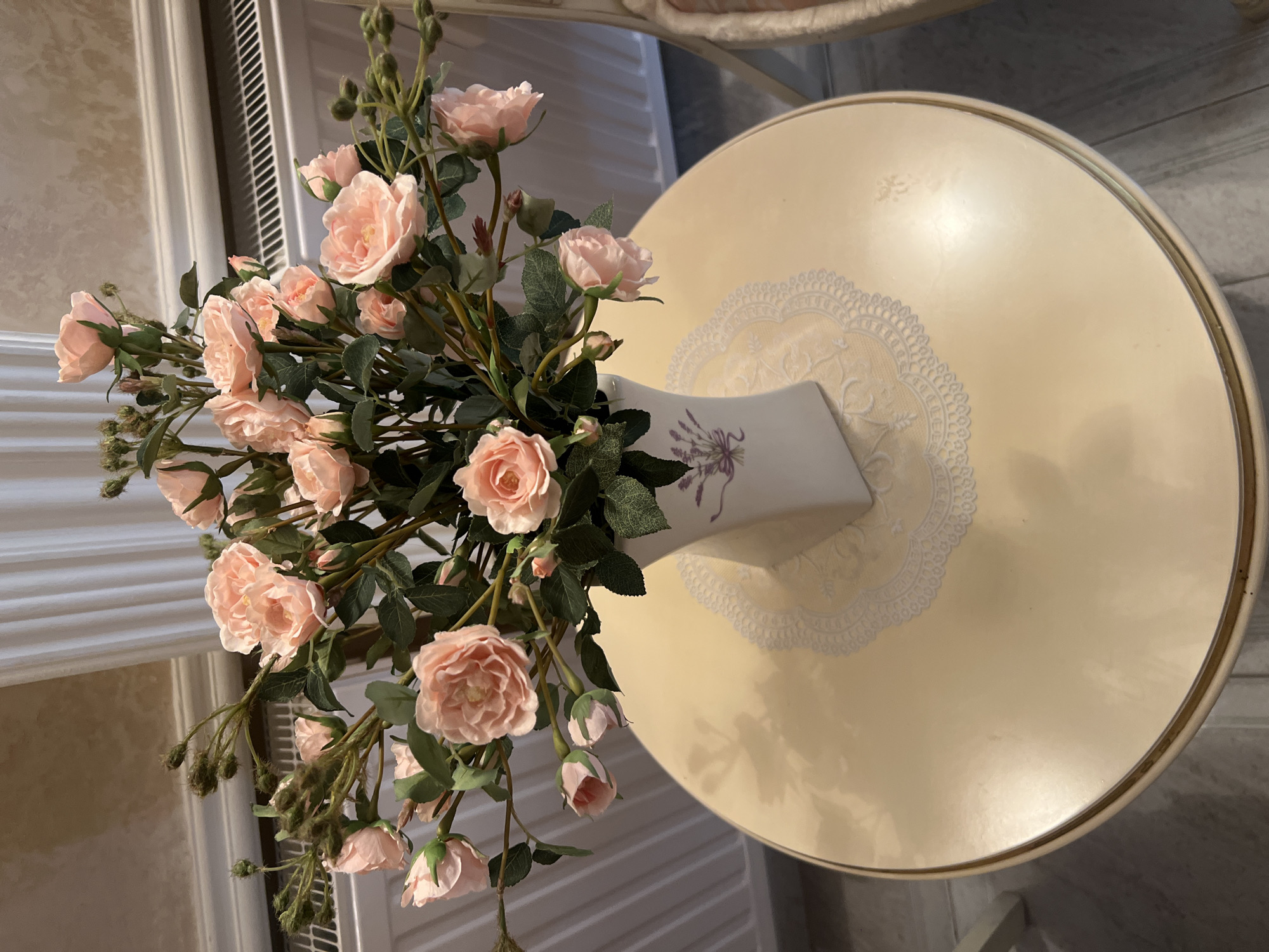 A vase of artificial pink roses sits on a cream-colored round table. The vase is white and decorated with a lavender sprig. The table has a doily underneath the vase. In the background, a white radiator and a floor are visible. The scene appears to be indoors, possibly in a home or a hotel in Bender, Moldova.