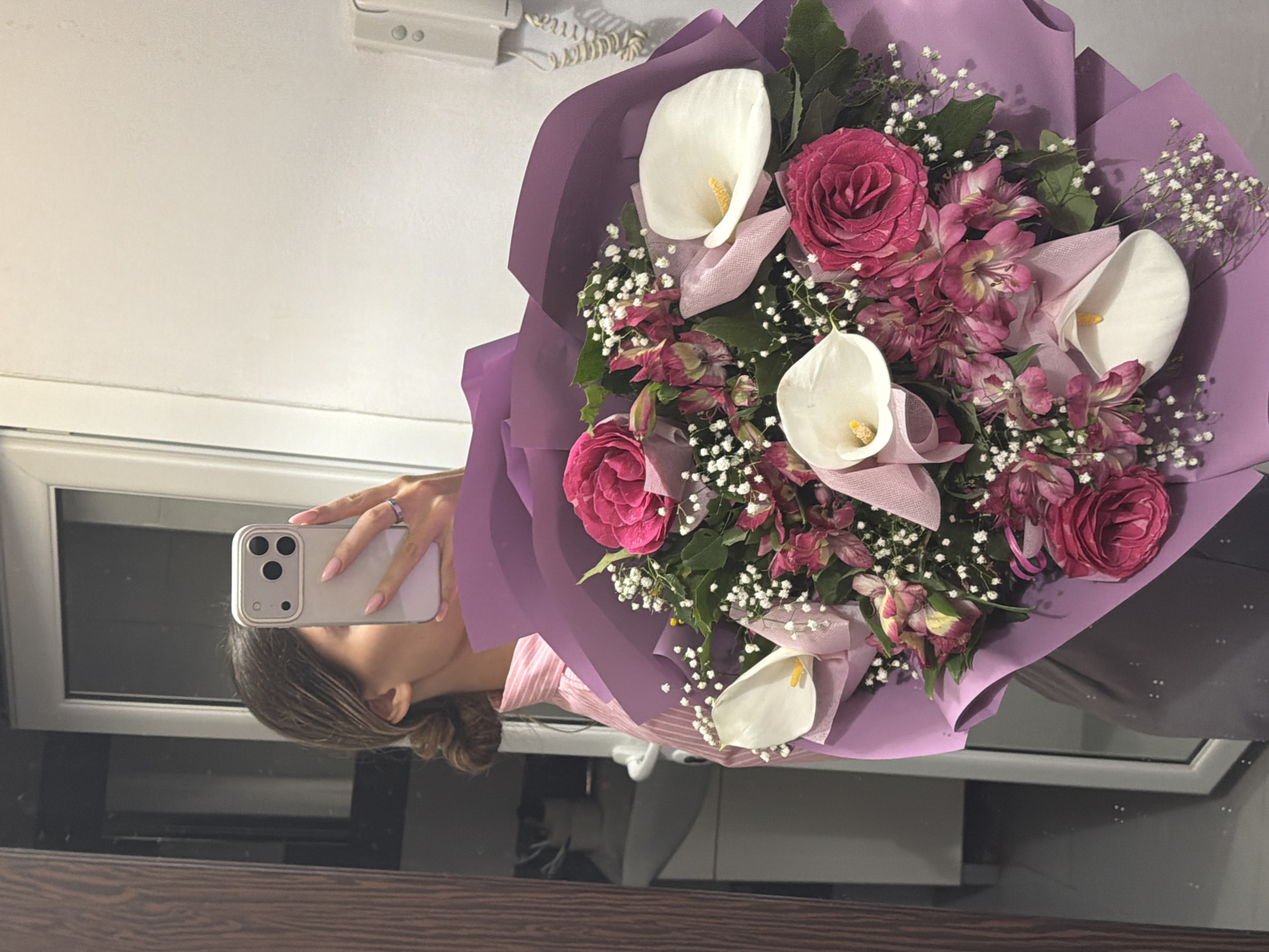 A person is taking a selfie in a mirror while holding a bouquet of flowers. The bouquet consists of white calla lilies, pink roses, and baby's breath, wrapped in purple paper. The person is holding a white smartphone with a visible camera array. A light-colored wall and a window frame are visible in the background, reflected in the mirror. The location in the city of Sofia, Bulgaria cannot be determined from the provided image.