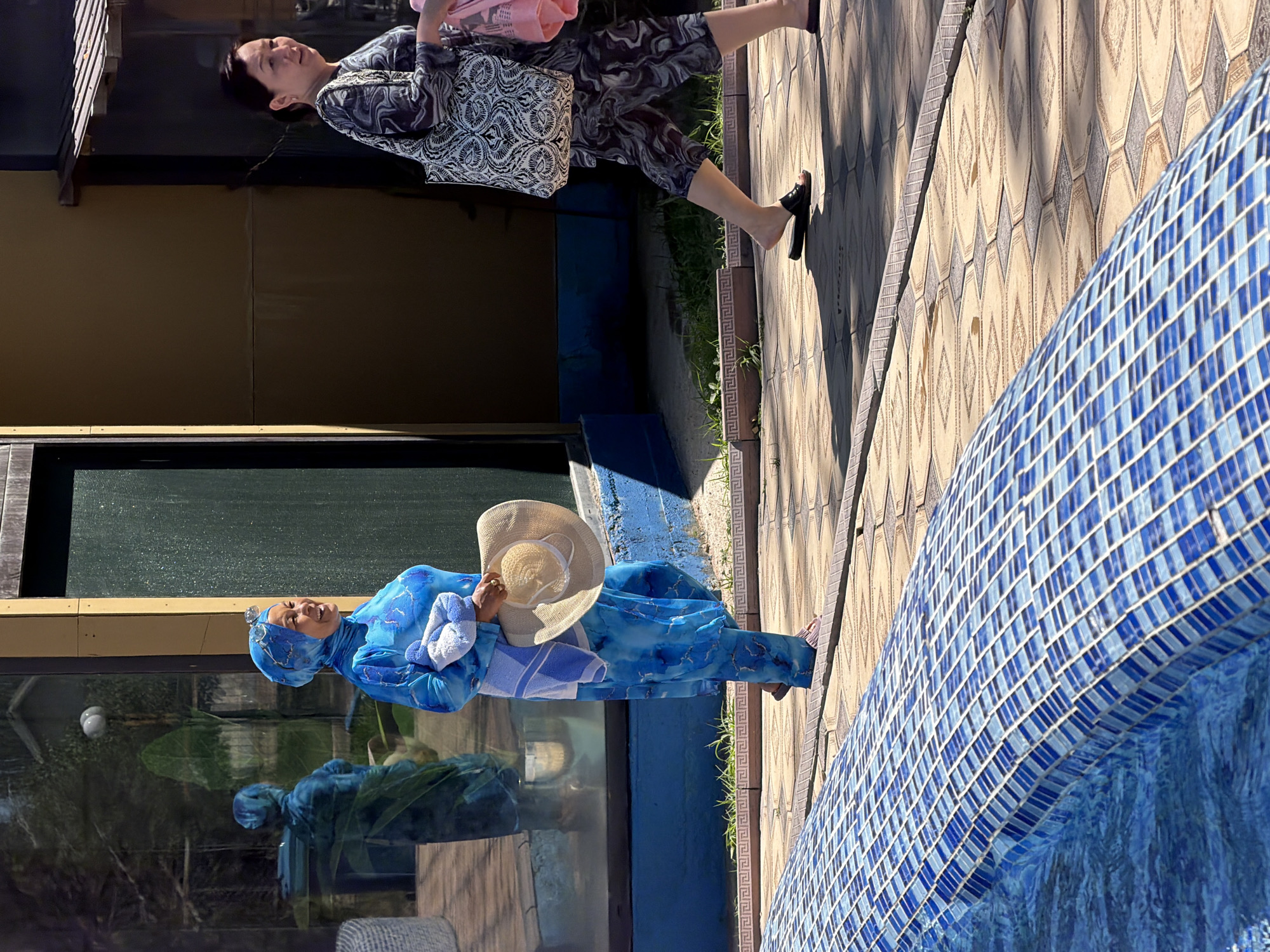 In this high-angle, full-shot image taken outdoors, two women are walking on a tiled path next to a blue-tiled swimming pool. The woman on the left is wearing a bright blue, patterned long-sleeved shirt and pants, a blue headscarf, and sunglasses on her head. She is holding a light-colored straw hat and a folded blue and white towel in her arms. She is smiling and looking to her right. The woman on the right is wearing a dark, patterned long-sleeved shirt and pants. She is carrying a large tote bag with a black and white pattern on her left shoulder and a pink bag in her right hand. She is looking to her left and walking away from the camera. The ground is paved with light-colored square tiles arranged in a diamond pattern with darker geometric accents. The swimming pool on the left is tiled with small blue and white squares. In the background, a building with a yellow facade and dark green doors is visible, along with reflections in its windows. Sunlight casts shadows on the tiled path.