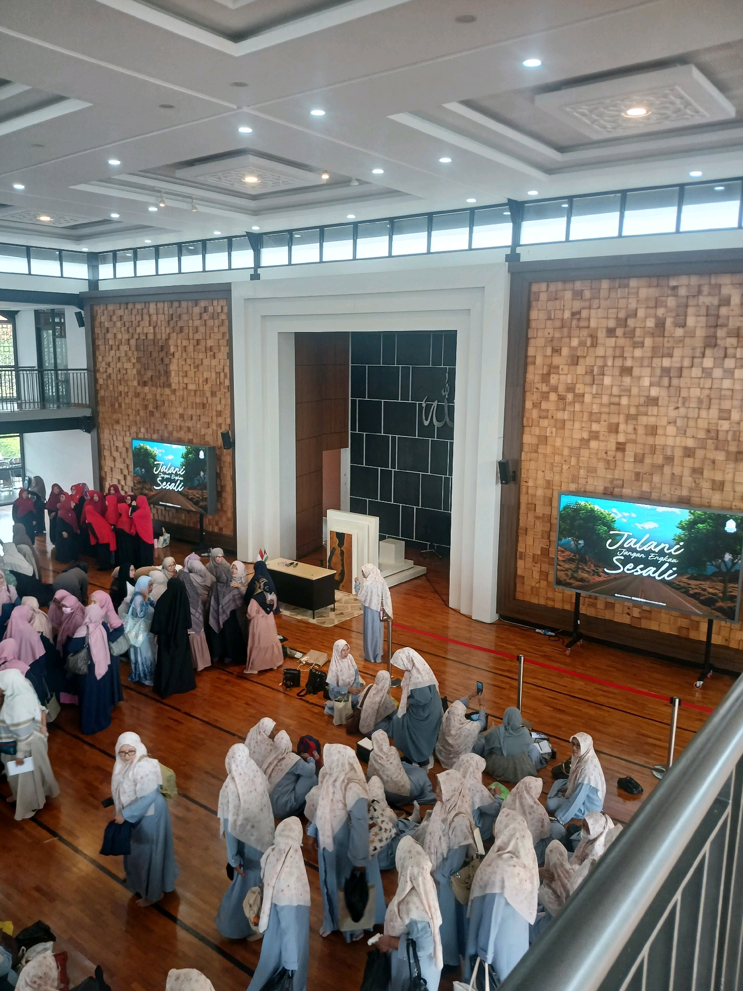 The image shows a large indoor gathering of Muslim women, predominantly dressed in hijabs and modest clothing, in a hall with wooden walls and flooring. Two large screens display the text "Jalani Dengan Bersuci" which translates to "Live with Purity". One screen is positioned on the left side of the hall, and the other on the right. In the center of the hall, there is a raised platform with a podium and some chairs, suggesting a presentation or speech is taking place. The women are seen standing and sitting in groups, some interacting with each other, while others appear to be listening. The lighting in the hall is a mix of ambient and spotlight illumination, creating a bright and spacious atmosphere. The overall scene suggests a religious or educational event. Given the context of Arjasari, Indonesia, this could be a mosque, a community center, or an educational institution.