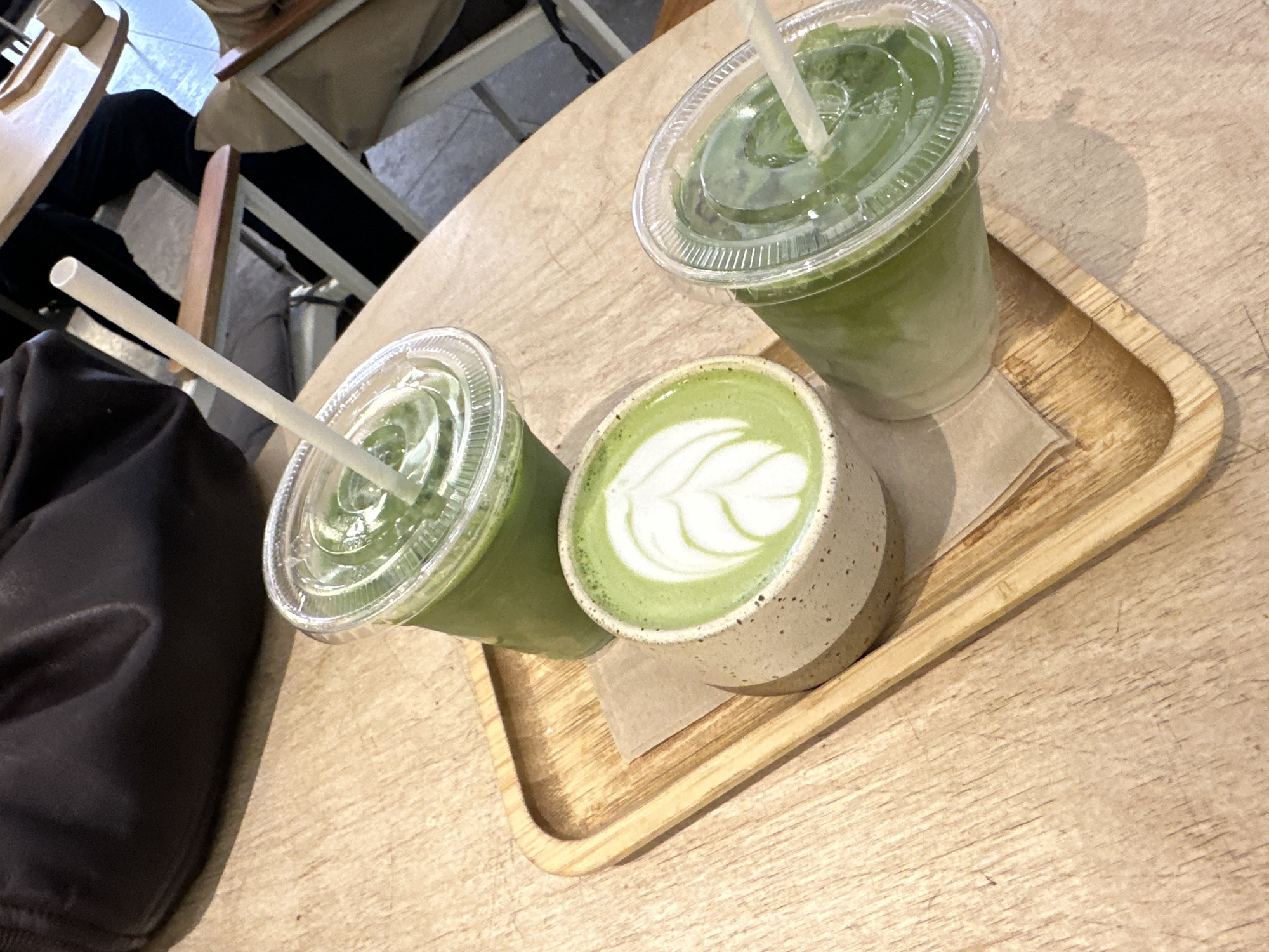 The image shows a wooden tray on a wooden table holding two plastic cups of matcha drinks with lids and straws, and a ceramic cup of matcha latte with latte art. The scene appears to be indoors, possibly a café. Part of a person and a chair are visible in the background. The location is unknown.