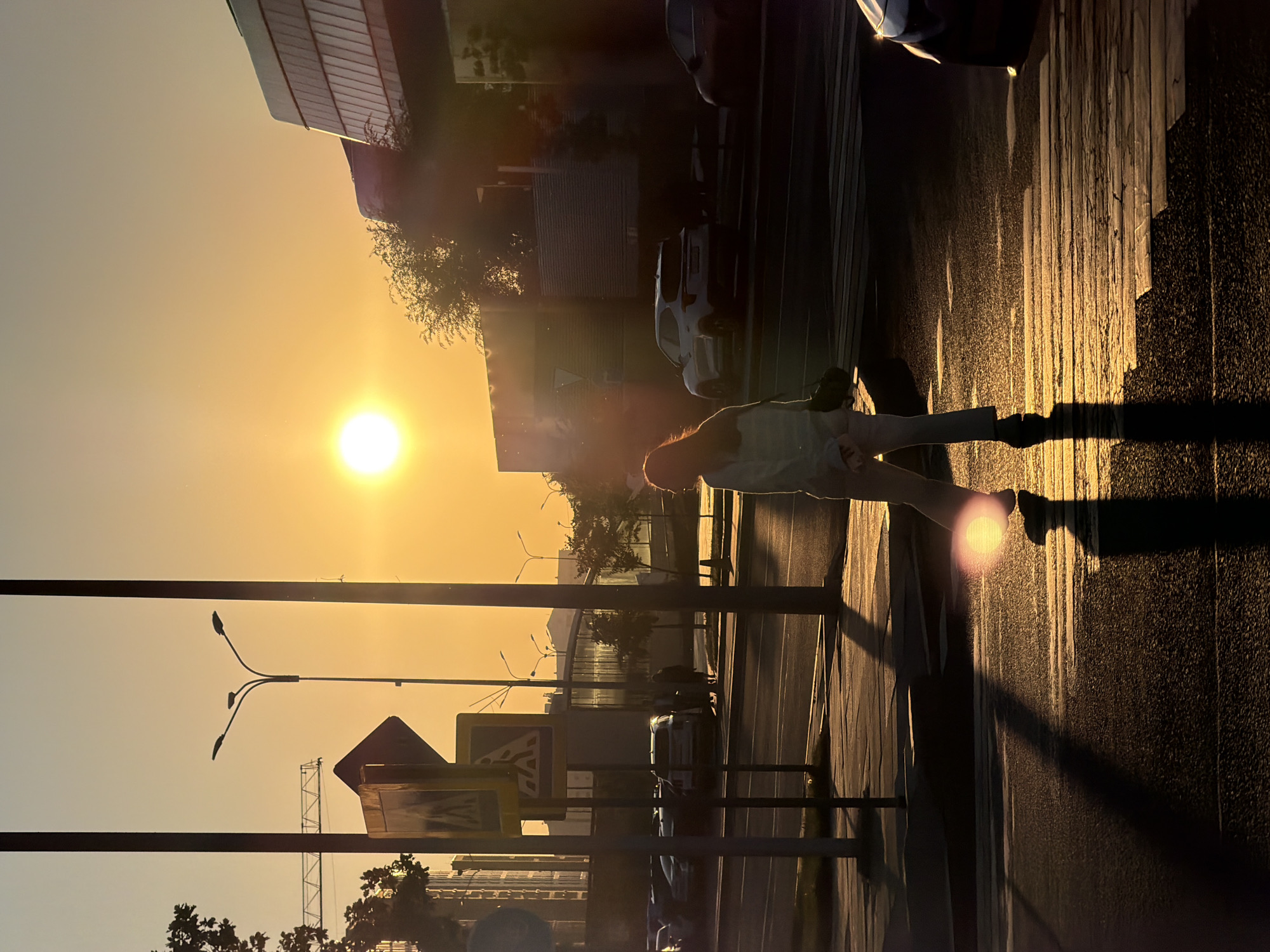 The image depicts a city street scene in Astana, Kazakhstan, during sunset. The low-angled sunlight casts long shadows across the asphalt and a pedestrian crossing. A single person, silhouetted against the bright sky, is walking across the street. The individual appears to be wearing light-colored pants and a jacket, with dark hair. In the background, several cars are parked, and modern buildings with glass facades rise above the trees. Streetlights and traffic signs are visible along the roadside. The overall atmosphere is serene, illuminated by the warm, golden hues of the setting sun.