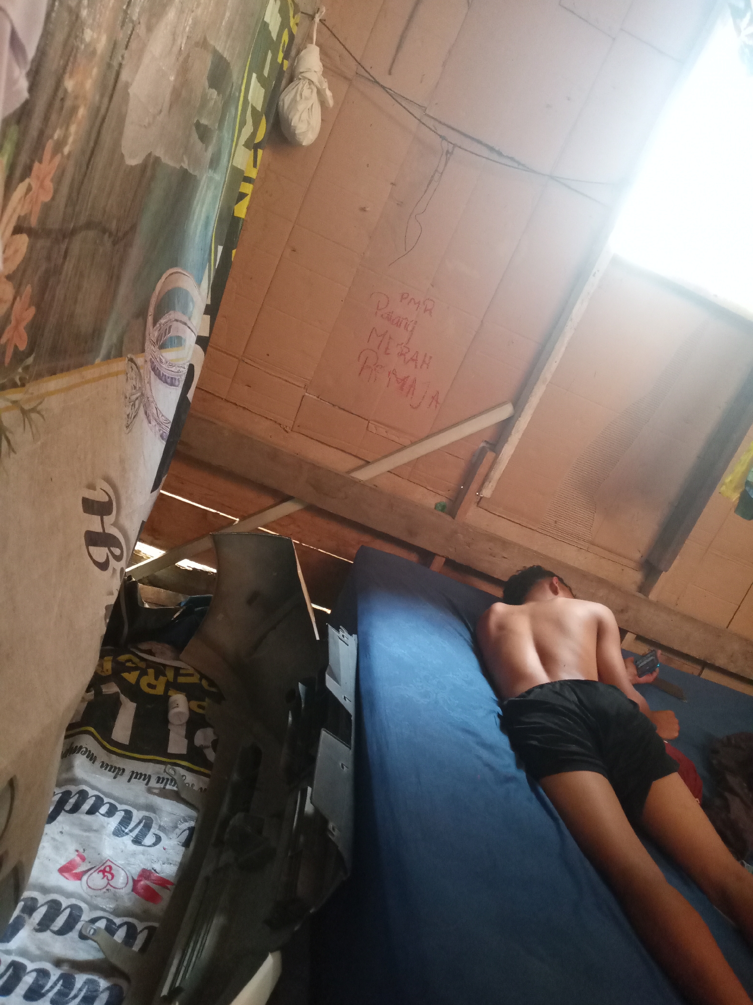 A young male is lying on his back on a blue mattress. He is wearing black shorts and his torso is bare. He is holding a black smartphone in his right hand. The man is in a dimly lit room with a wooden ceiling. There are handwritten words in red on the ceiling which appear to say "PMR Padang MERAH PEMUDA". A bundle wrapped in white paper is hanging from a string attached to the ceiling. The room is cluttered with various items including what appears to be a mattress leaning against a wall. The overall environment suggests a makeshift or storage space. The location is Unknown, Unknown.