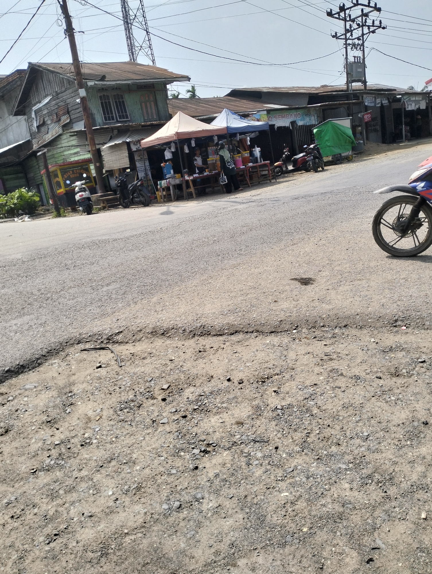 The image depicts a street scene in Pematang Lumut, Indonesia. The foreground is dominated by a rough, gravelly road surface with some visible wear and tear, including a depression and some scattered debris. In the midground, a row of buildings lines the street, appearing to be shops or small businesses. These structures have weathered exteriors, with some appearing to be made of wood and corrugated metal. Several market stalls or temporary structures are set up in front of the buildings, some covered by awnings.
Various objects are present in the scene. Two motorcycles are parked near the stalls. One stall has a sign with "Q SEMBAKO" visible. Another stall has a yellow sign with "FRIED CHICKEN FOOD" in English. In the background, a tall electrical transmission tower stands against a hazy sky. A portion of a motorcycle, specifically its front wheel and fender, is visible in the right foreground, suggesting a vehicle is either passing by or parked close to the camera. The overall impression is one of a bustling but somewhat dilapidated commercial area in a tropical or sub-tropical setting, characterized by its unpaved road and modest infrastructure.