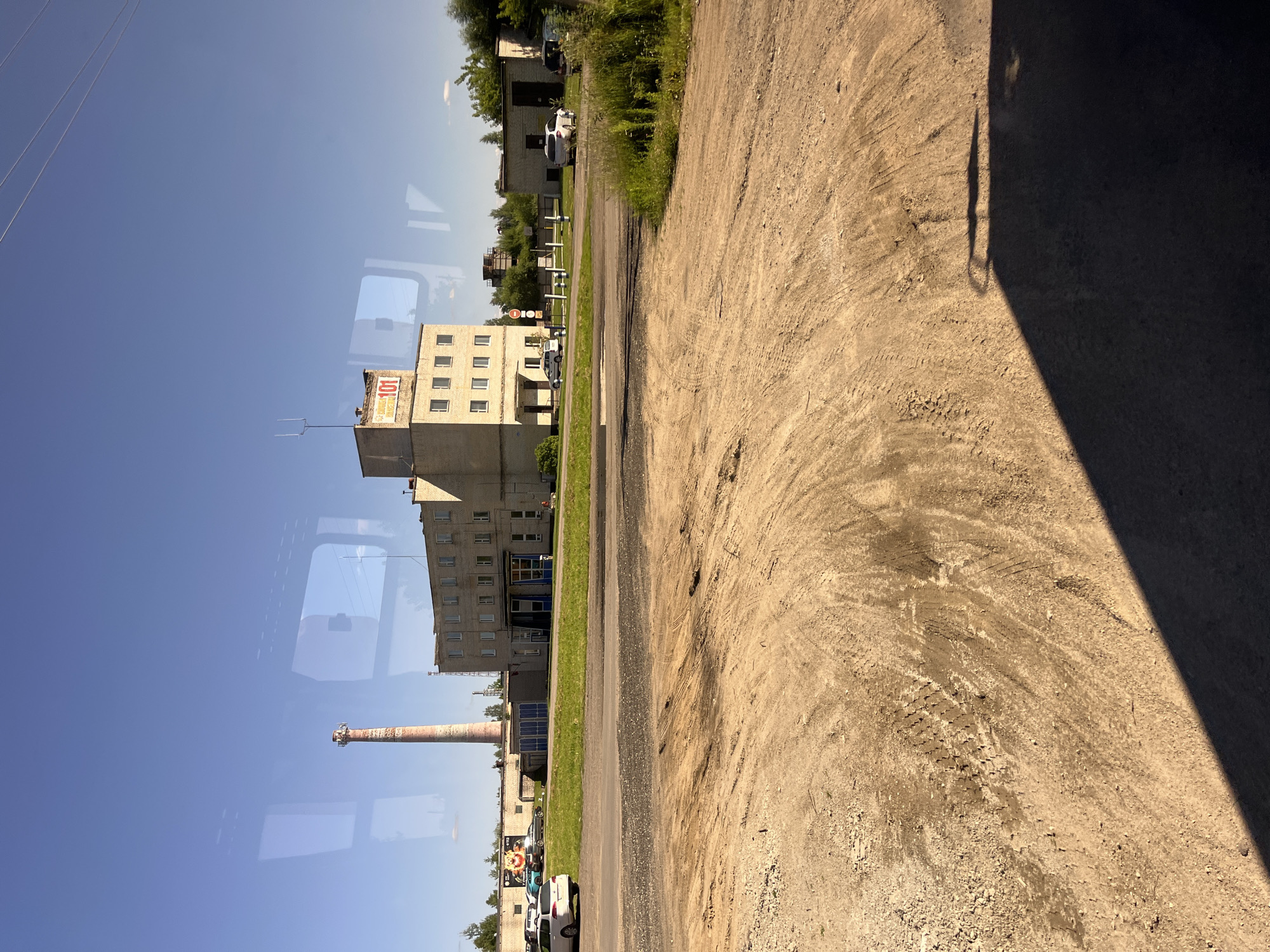 The image captures a view of an industrial area in Pastavy, Belarus, from what appears to be inside a vehicle due to reflections on the glass. In the background, a tall, red and white striped smokestack stands prominently, a characteristic landmark of the city. Adjacent to it is a light-colored brick building with multiple windows and a prominent sign on its roof displaying the number "101" in red letters. Other low-lying industrial buildings and structures are visible, along with trees and greenery. In the foreground, a rough, unpaved area with tire tracks is present, suggesting it's a service road or parking area. Several vehicles, including cars and a van, are parked in the background. The sky is clear and blue, indicating a sunny day. The overall scene depicts a functional, albeit somewhat dated, industrial complex within the city.