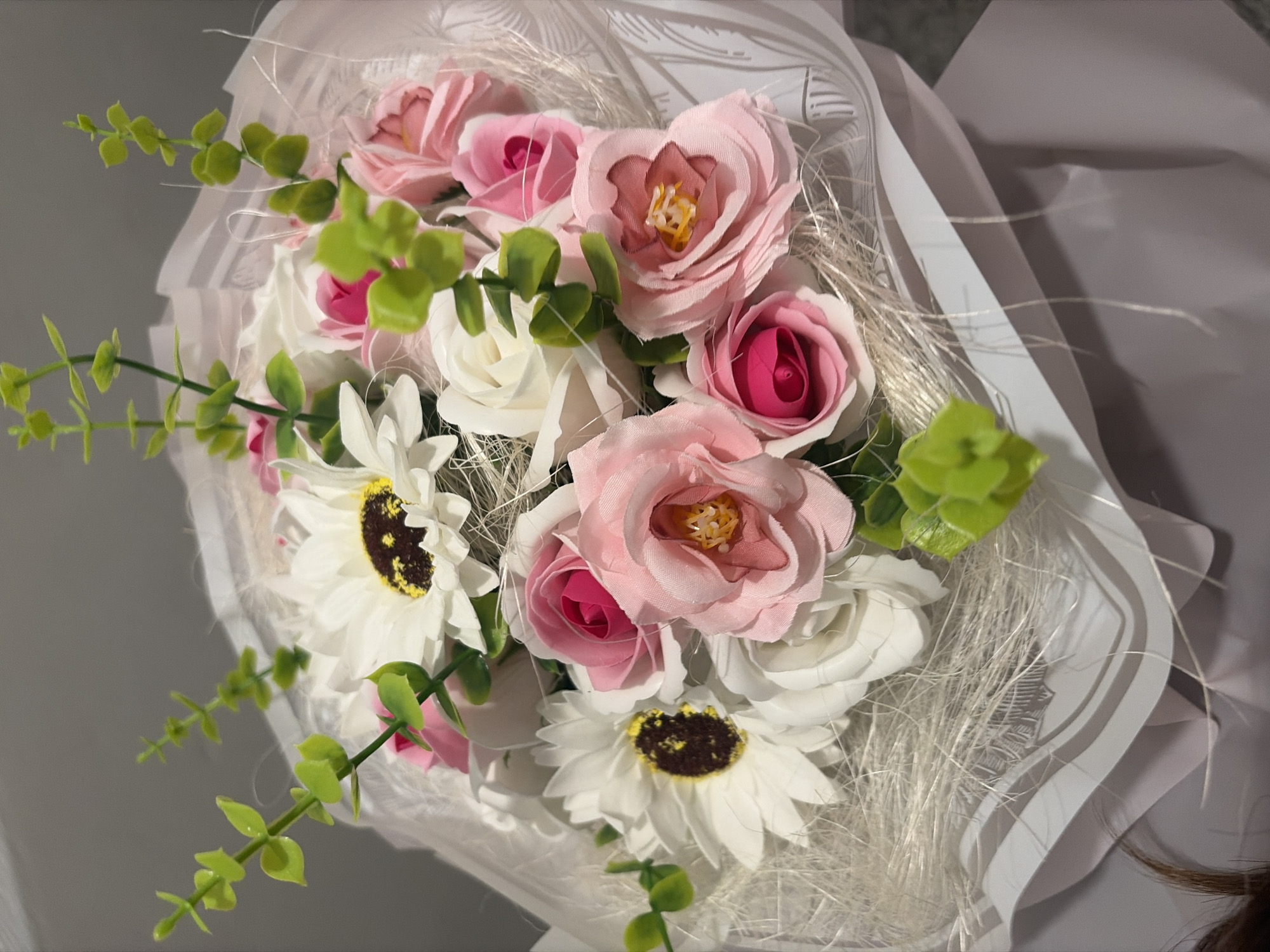 The image shows a bouquet of artificial flowers in a white wrapping. The bouquet includes pink and white roses and white daisies with brown centers. Green leafy sprigs are interspersed among the flowers. Fine white fibers add a textured element to the arrangement. There is no indication of location, people, actions, or interactions within the image. The environment is indistinct and provides no location-specific information.