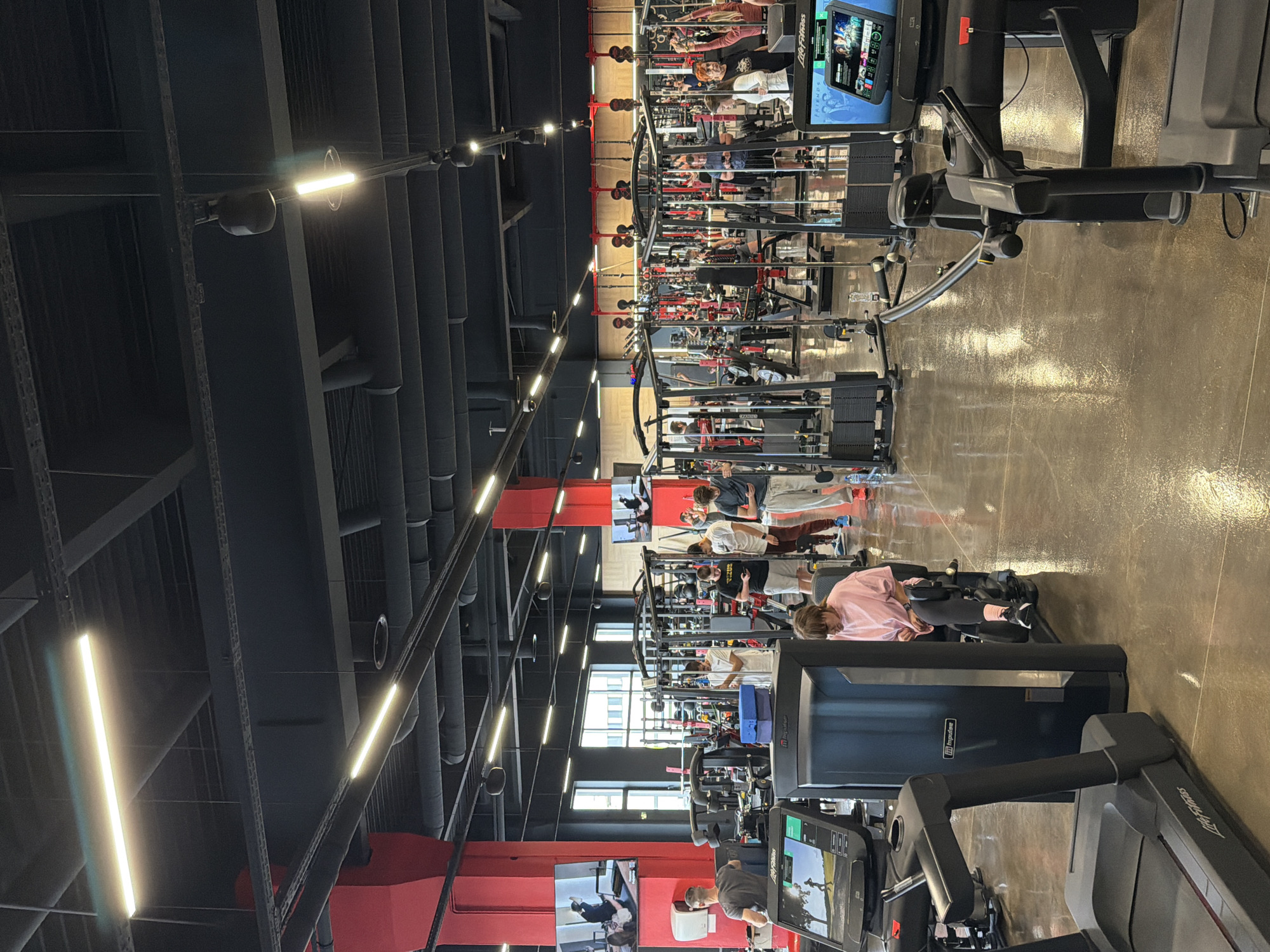 The image shows the interior of a gym in Plovdiv, Bulgaria. The gym has a dark gray ceiling with exposed pipes and industrial lighting. Red support pillars are visible. The floor is a polished concrete or epoxy. Various exercise machines are arranged in rows, including treadmills and weightlifting equipment. Several people are using the equipment, wearing workout clothes. The gym has large windows. There are television screens mounted on the walls, displaying content. The gym is actively in use.