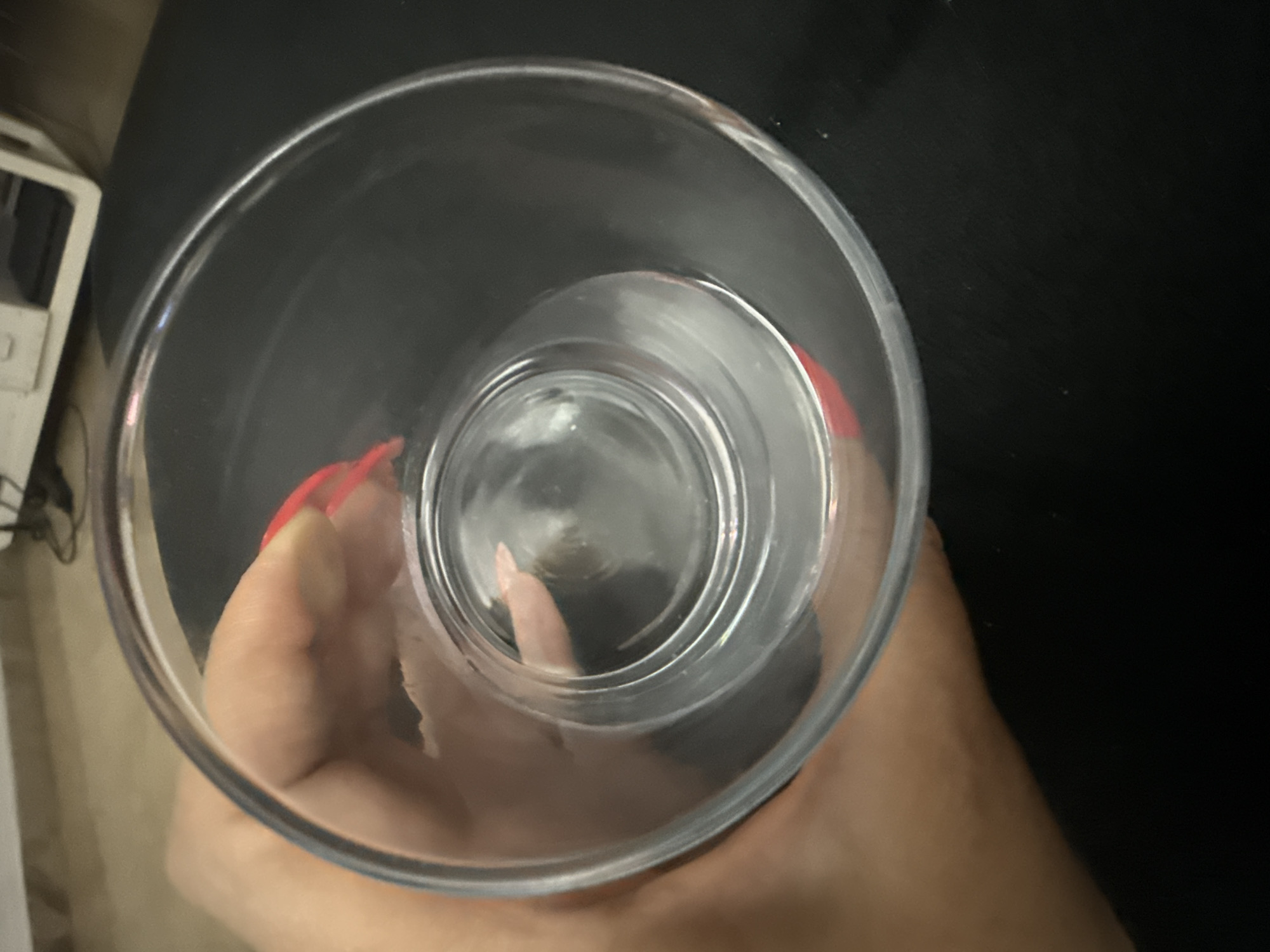 An overhead shot reveals a clear glass held by a hand. The glass appears to be empty or nearly empty, with light reflecting off the interior surfaces. The hand has polished nails. The background is a mix of dark and light surfaces, with a glimpse of what appears to be a white electronic device in the periphery. There is no indication of the location being Valencia, Spain.