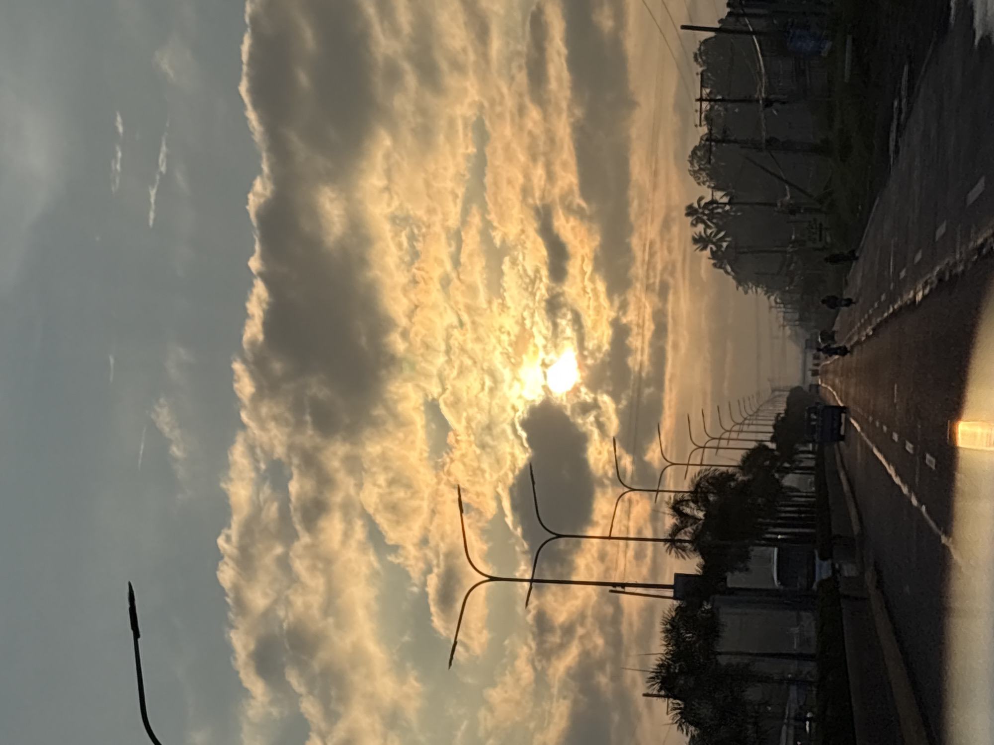 This media file captures a road scene in Carmen, Philippines, at what appears to be sunrise or sunset due to the warm, golden light illuminating the clouds. The sky is filled with dramatic, textured clouds, some of which are brightly lit by the sun, creating a beautiful, almost ethereal effect.
The scene depicts a road with multiple lanes, bordered by a sidewalk or shoulder. Along the side of the road, there is a row of streetlights with distinctive, multi-pronged designs. Further beyond the streetlights, palm trees and other lush vegetation are visible, suggesting a tropical or subtropical environment. There are a few vehicles on the road, including what appears to be a truck in the distance, and several motorcycles or smaller vehicles further down the road. The road surface seems wet, possibly indicating recent rain or morning dew.
The overall mood of the image is serene and peaceful, enhanced by the soft, warm lighting and the natural beauty of the sky and landscape. The time of day is suggested by the low angle of the sun and the dramatic cloud formations. While no specific text is clearly legible in the image, the presence of streetlights and the paved road indicate an established infrastructure, likely a highway or a significant thoroughfare within the Carmen area.