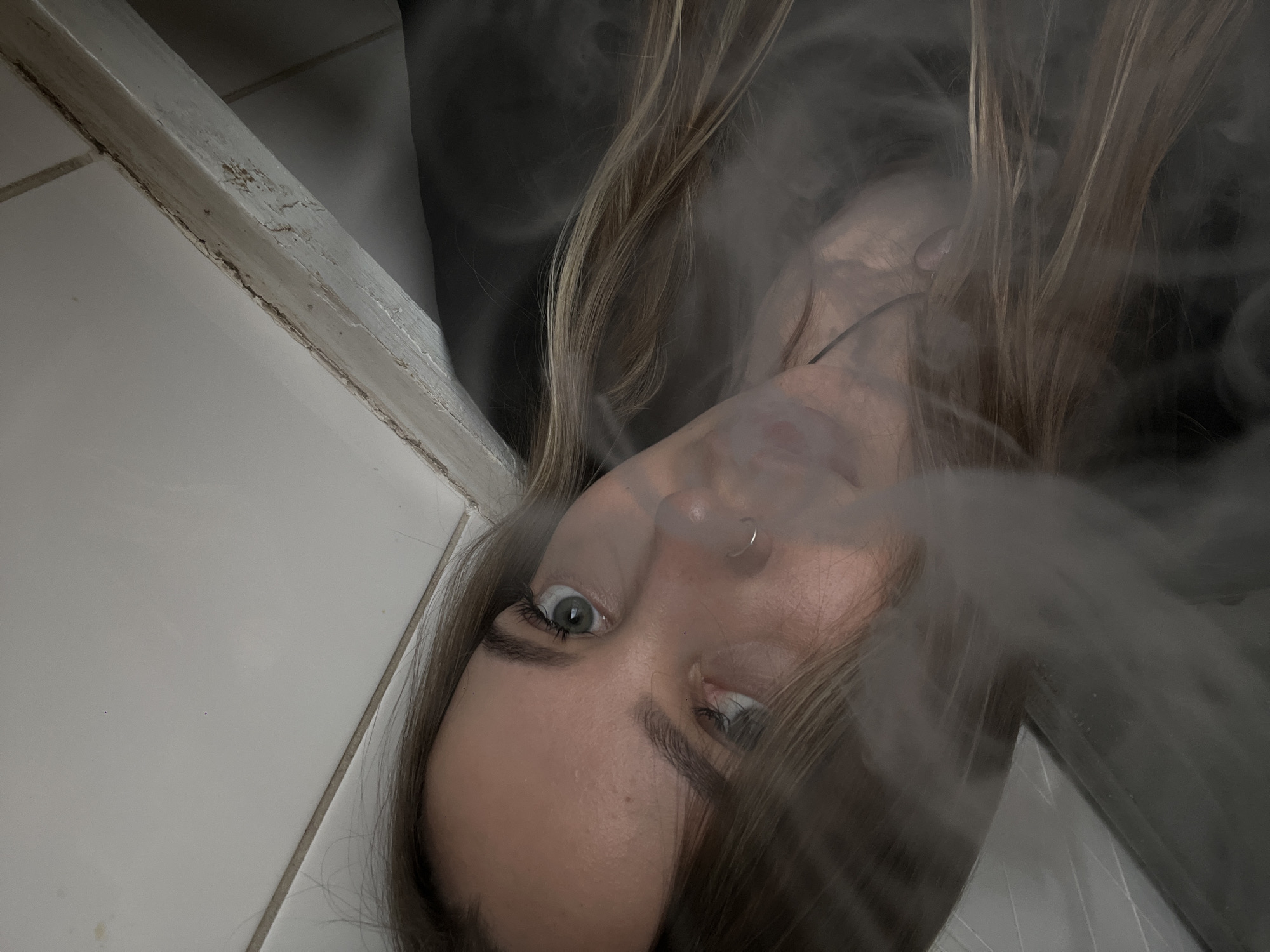 A woman with light brown hair and blue eyes is positioned in the center of the frame, surrounded by smoke. She has a nose ring in her left nostril and is exhaling smoke from her mouth. The background includes white tiled walls, with a trim visible along the top edge of the tiles. A dark, blurry area is visible in the upper part of the frame, above the tiled wall. The location is not discernible from the image, so providing the location in Novopokrovskaya, Russia is not possible based on the visual information available.