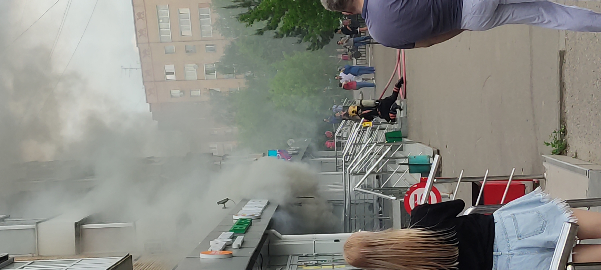 The image shows a building in Unknown, Unknown, obscured by heavy smoke emanating from the front of the structure. A crowd of onlookers stands behind a metal railing observing the scene. A firefighter in full gear is positioned near the entrance, presumably responding to the fire. In the foreground, part of a person is visible, wearing a denim skirt and a black top, and another is visible wearing white pants and a grey top. The building appears to be a commercial establishment, identifiable by signage on its facade. The sky is overcast.