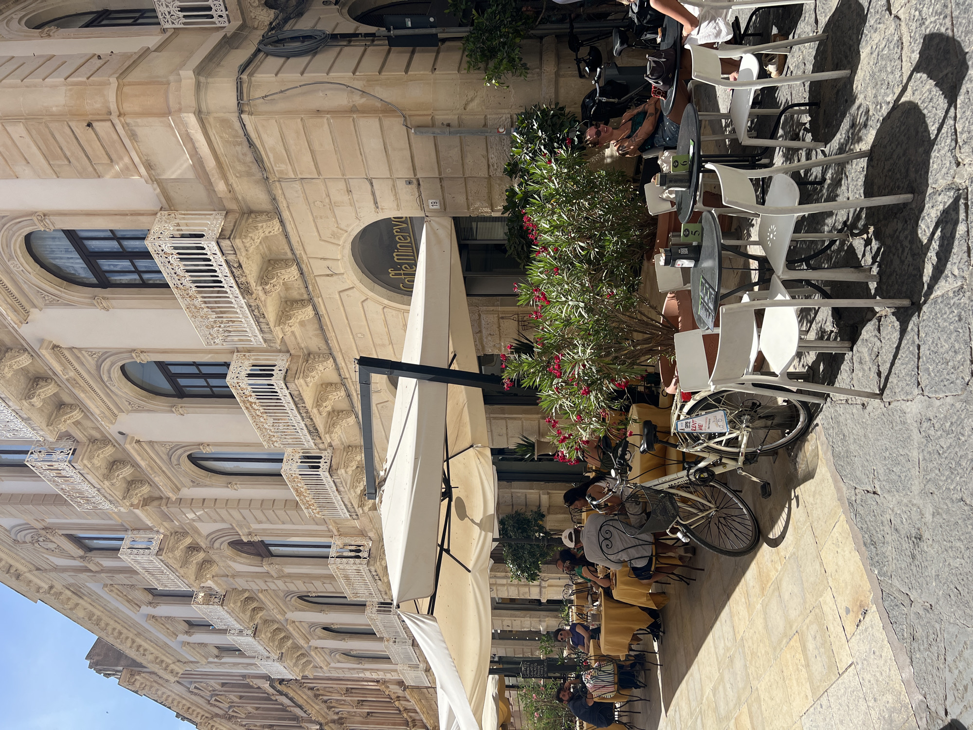 In the city of Syracuse, Italy, an outdoor cafe is depicted on a cobblestone street. The cafe has several tables and chairs set up under large white umbrellas. People are seated at the tables, some eating and drinking, while others are engaged in conversation. A white bicycle with a basket is parked near one of the tables. The buildings surrounding the cafe are made of light-colored stone and feature ornate balconies. Lush green plants with red flowers add a touch of color to the scene. The sun is shining, casting shadows on the ground. The cafe is identified as "Caffè Minerva" by a sign above the entrance.