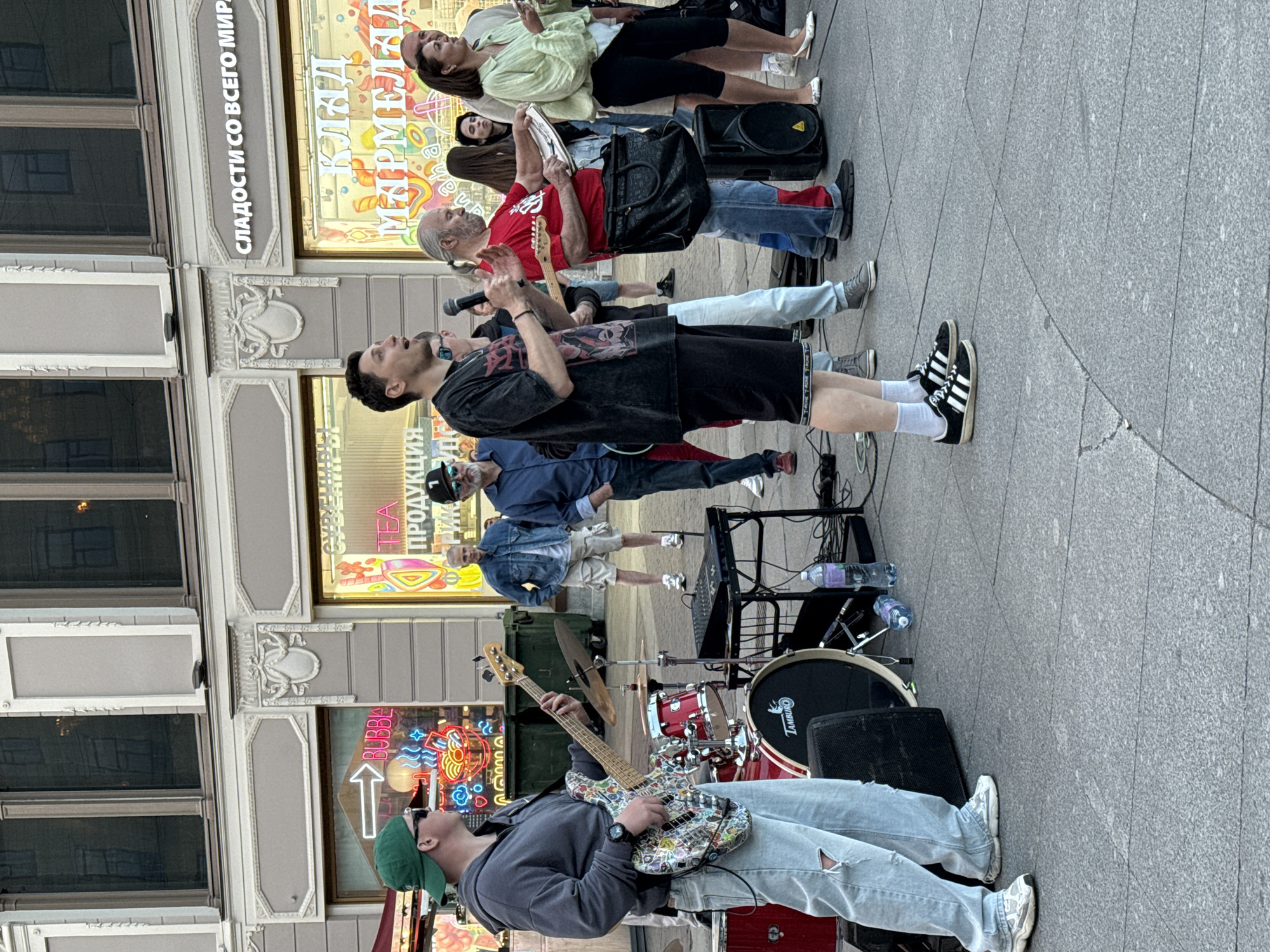 The image captures a street performance in Saint Petersburg, Russia, likely on Nevsky Prospekt, given the architecture and storefronts. A band is set up on the pavement, with a bassist in the foreground playing a sticker-covered bass guitar. A singer stands next to him, holding a microphone and facing the audience. Behind them, a drummer is partially visible with a drum kit. Several other band members and onlookers are also present, some interacting with the performers. The background features storefronts with signage in Russian, including "СЛАДОСТИ СО ВСЕГО МИРА" (Sweets from all over the world) and "КЛАД МАРМЕЛАДА" (Treasure of Marmalade), suggesting a commercial area. The overall scene depicts a lively street event in a bustling urban environment.