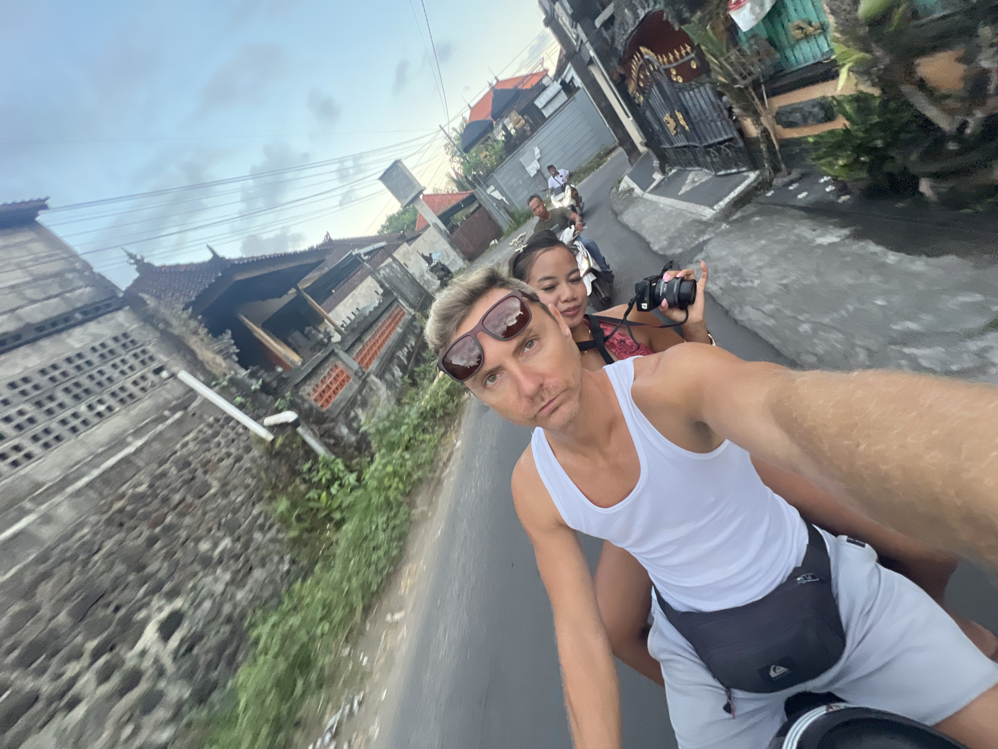 The image shows a man in a white tank top and sunglasses riding a motorbike in the city of Sukawati, Indonesia. A woman is behind him, holding a camera. Other people on motorbikes are further ahead on the road. The surrounding environment consists of buildings with traditional Indonesian architectural elements and lush vegetation along the roadside. The road is paved, and the sky appears overcast. The man is taking a selfie while driving.