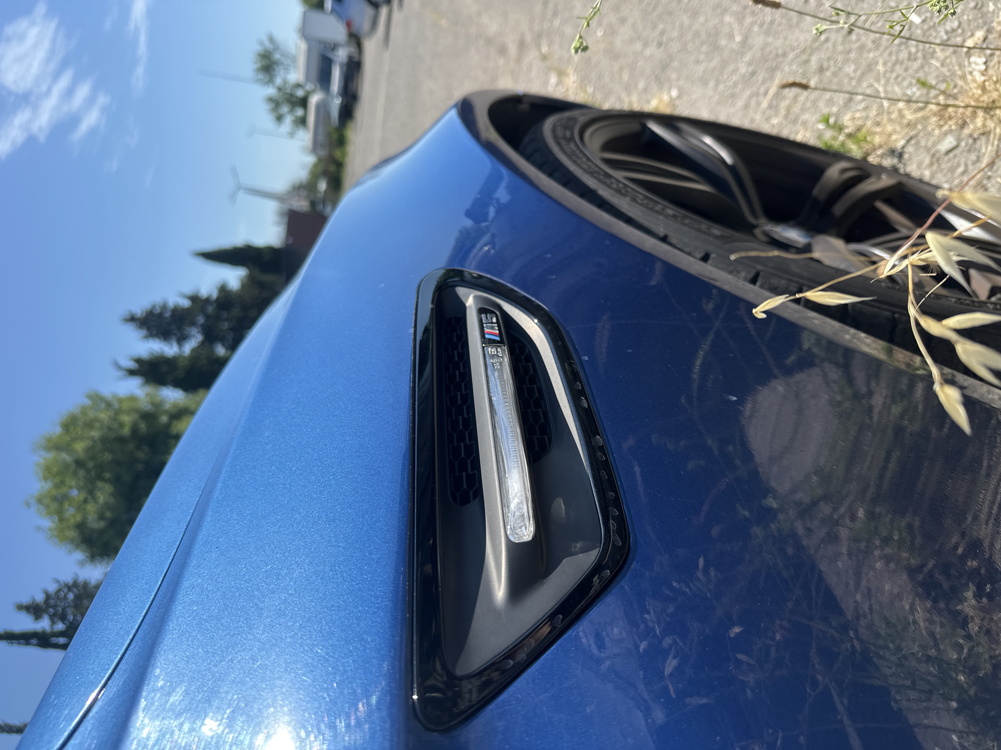 A blue BMW with an M badge is parked outdoors in a lot. The front fender and wheel are visible, along with a portion of the vehicle's hood. The car is angled towards the right, and the background shows a clear blue sky with some clouds, trees, and other parked vehicles. The ground is paved and dry, with some weeds growing near the car's tire.