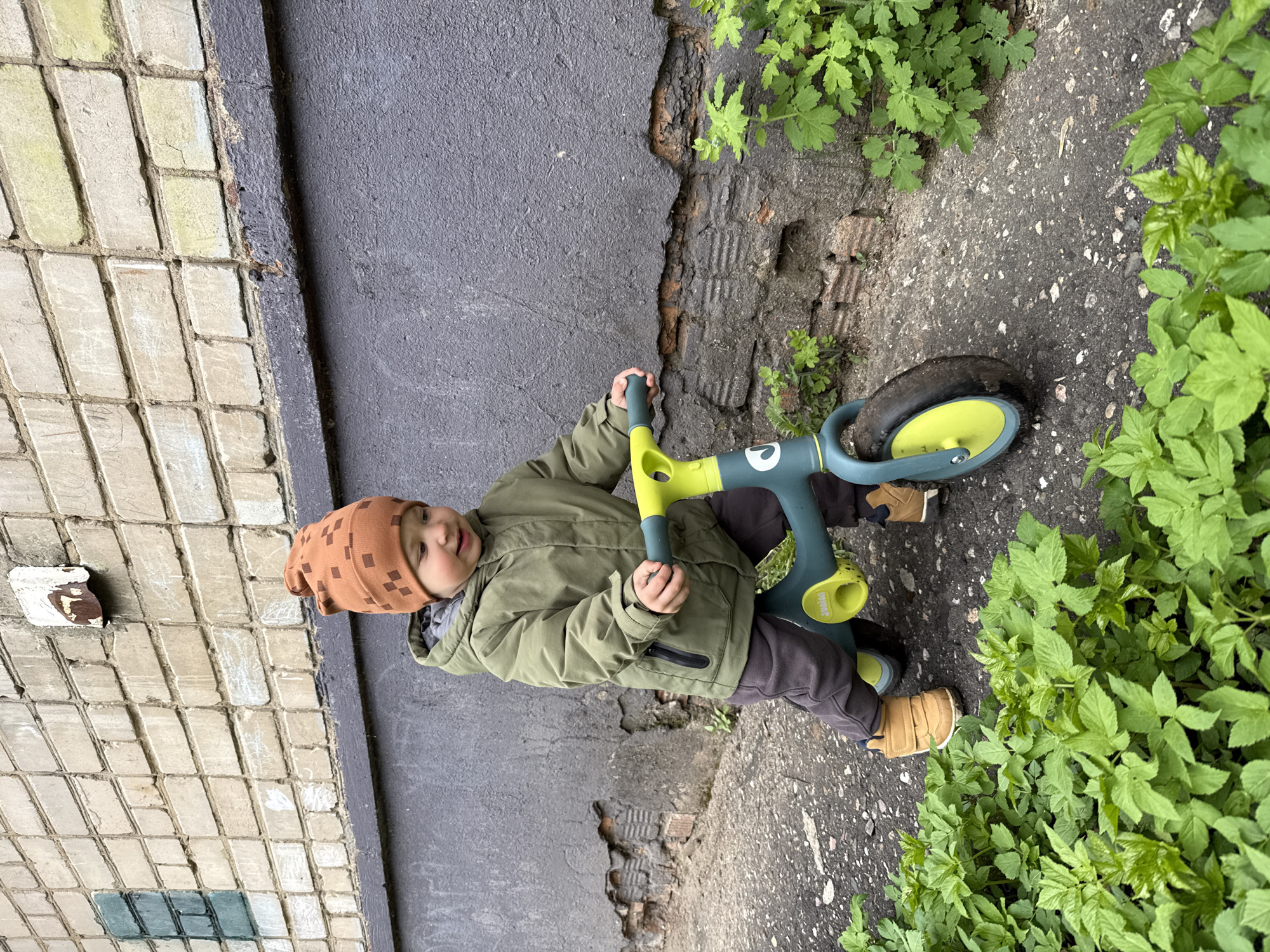 A child sits on a toy tricycle on a paved surface, positioned next to a wall. The wall is comprised of two distinct sections: one side is a black, textured surface and the other is covered with light-colored, rectangular tiles with some green staining. Part of the textured surface is cracked. Green leafy plants grow along the edge of the paved surface, adjacent to the tricycle's rear wheel. The child, wearing a brown patterned hat, an olive green jacket, brown pants, and tan shoes, is gripping the tricycle's handlebars.