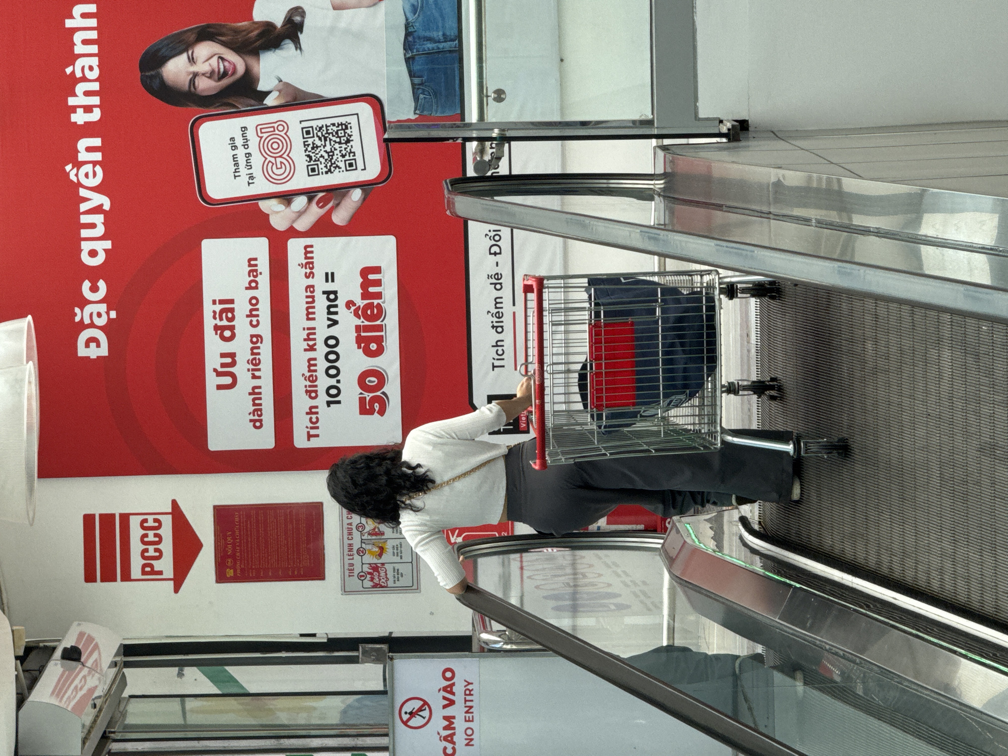 The image shows a woman with curly dark hair, wearing a white cropped long-sleeved top and gray pants, standing on an escalator. She is pushing a shopping cart with a red and blue bag inside. To her left is a sign that reads "CẤM VÀO NO ENTRY" with a red circle and a figure walking. Above the escalator, a red banner displays Vietnamese text and a QR code on a smartphone. The scene appears to be an indoor shopping environment, possibly a supermarket or department store, with a modern design. The presence of the escalator and shopping cart suggests a commercial setting. The red banner with Vietnamese text and a QR code indicates a promotion or loyalty program. The "NO ENTRY" sign suggests a restricted area. The overall setting is clean and well-lit, typical of a retail space. The image is taken from a slightly low angle, looking up the escalator.