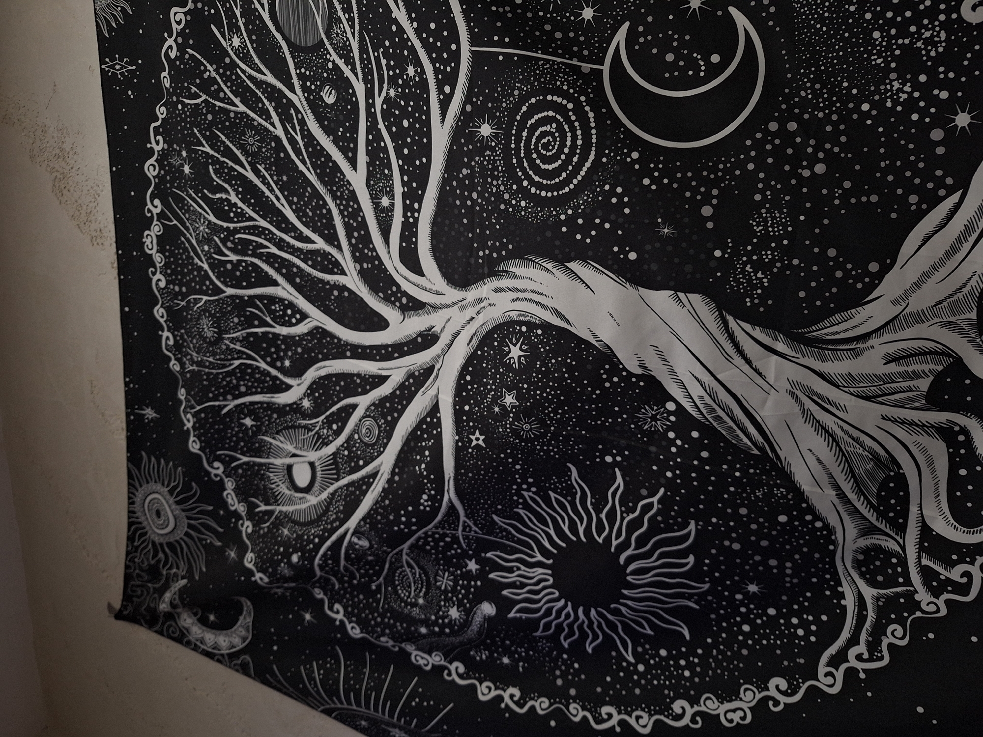 The image displays a large, black and white tapestry hanging on a light-colored wall in what appears to be an indoor setting in Panagyurishte, Bulgaria. The tapestry's central motif is a highly stylized, intricate tree, often referred to as a "Tree of Life," with numerous branching limbs and roots extending across the fabric.
The background of the tapestry is a deep black, peppered with countless white dots of varying sizes, simulating a starry night sky or a galaxy. Prominent celestial elements include a distinct white crescent moon positioned in the upper right area. Additionally, two radiant sun-like or starburst designs are visible towards the lower sections of the tapestry, one smaller in the lower left and a larger, more detailed one in the lower right.
The design incorporates other mystical and cosmic details such as spiraling patterns, smaller stylized stars, and an ornate, wavy border that frames portions of the artwork. The overall scene evokes a sense of cosmic wonder and mythical connection. No activity or event is taking place; the tapestry is displayed as decorative art. There is no visible text on the tapestry. The room is well-lit, highlighting the monochrome details of the artwork.