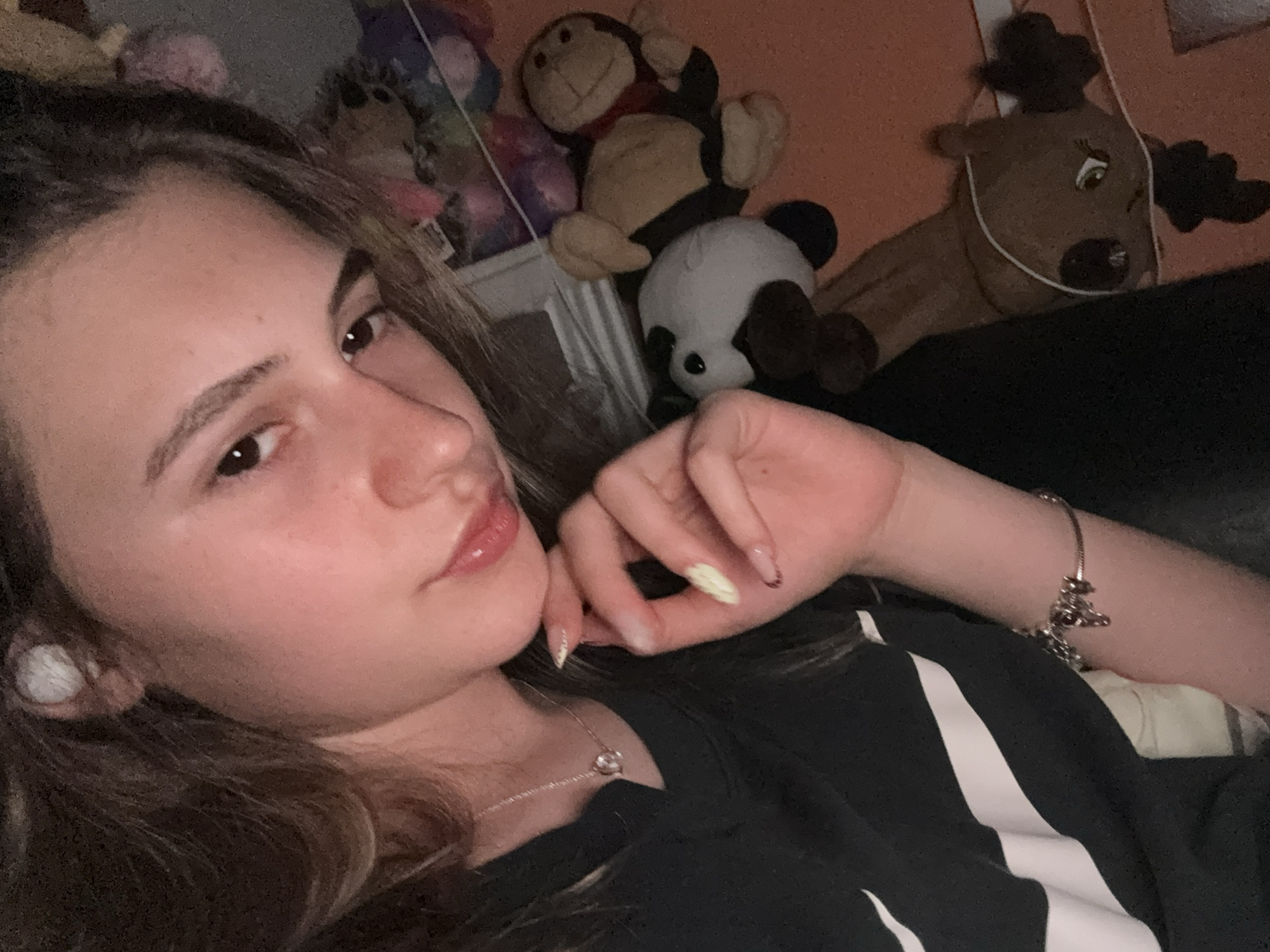 A close-up, head-and-shoulders shot shows a young woman with long, dark brown hair and fair skin. She is wearing a black t-shirt with a white diagonal stripe and a silver necklace with a circular pendant. She has white earbuds in her ears and light-colored nail polish on her fingers. Her right hand is near her face, and her left arm is visible with a charm bracelet. Behind her, several stuffed animals are visible, including a panda, a monkey, and a reindeer. The background appears to be a room with orange and gray surfaces, possibly a bed or couch. The overall lighting suggests an indoor setting, possibly at night or in a dimly lit room. There are no visible indications of the specific location within the city of Panagyurishte, Bulgaria.