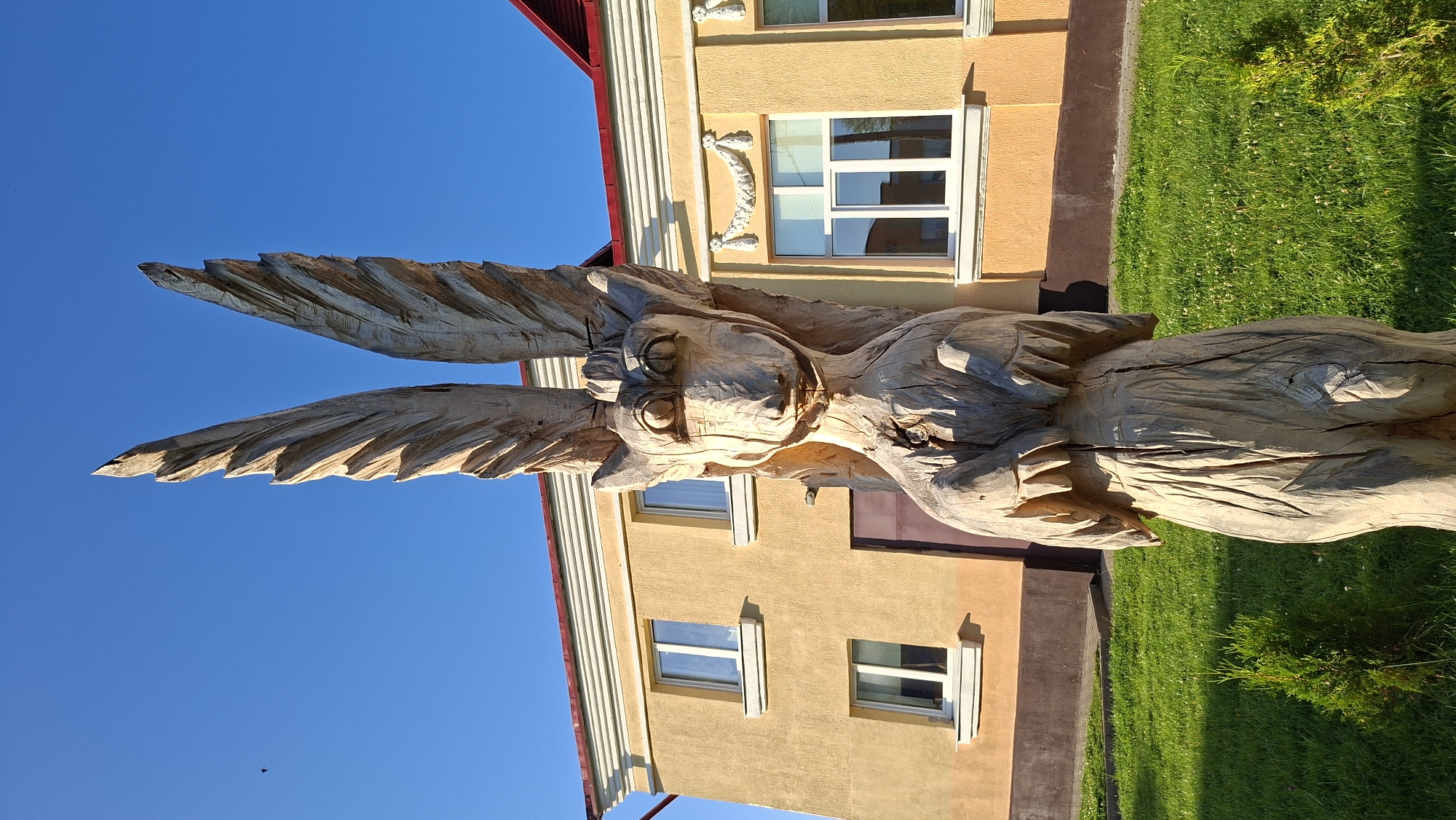 A wooden sculpture of a winged creature stands in front of a two-story yellow building with a red roof in Myadzyel, Belarus. The sculpture is carved from a light-colored wood and depicts a creature with large, feathered wings extending upwards, a distinct facial structure with prominent eyes and a snout, and what appear to be clasped hands or forelimbs. The building features several windows with white frames and a decorative trim above them. The ground in front of the building is covered in green grass, with a small evergreen bush visible to the left of the sculpture. The sky is clear and blue. A single bird is visible flying in the upper left corner of the image.
