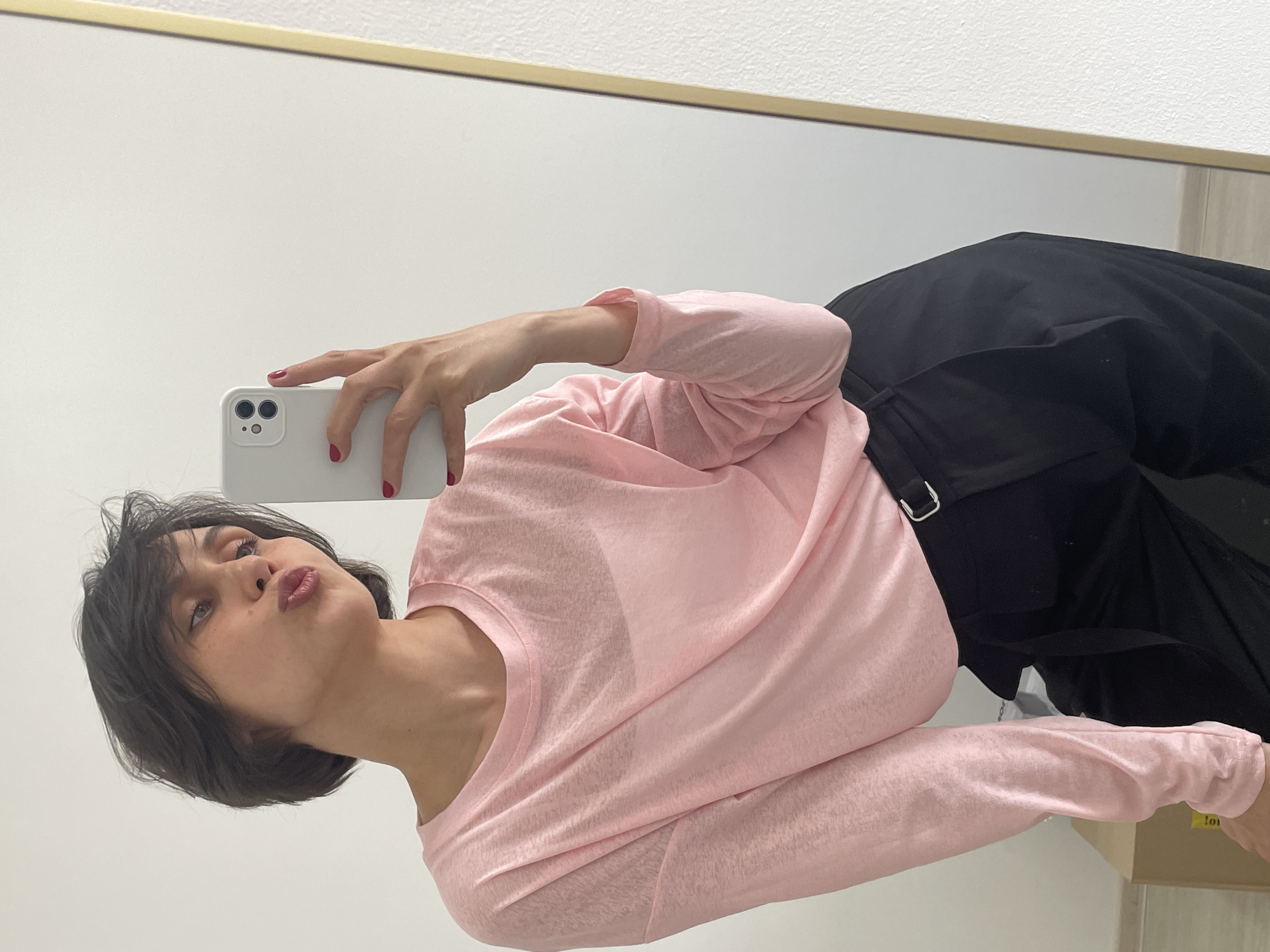 A woman with short, dark hair is taking a selfie in a mirror. She is wearing a light pink long-sleeved shirt and black pants. Her lips are pursed in a kiss. The mirror has a gold frame. The background is a white wall, with part of a beige wall visible on the right side. No information about the city of Izhevsk, Russia, can be discerned from the image.