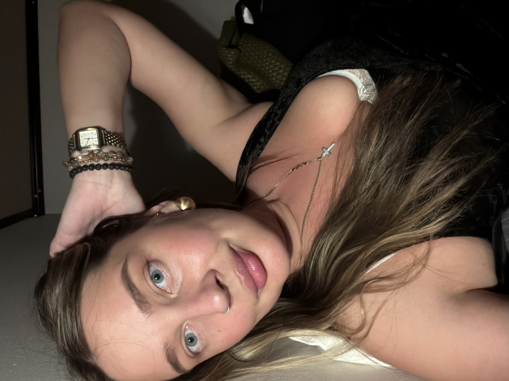 A woman lies on a light-colored surface. She is wearing a black top and has fair skin, blonde-brown hair, and blue eyes. She wears a watch and several bracelets on her left wrist. A gold necklace with a cross pendant adorns her neck. She has a nose ring and an earring in her left ear. A green woven bag is partially visible in the background. The location is not discernible from the image. The image does not show the city of Moscow, Russia.