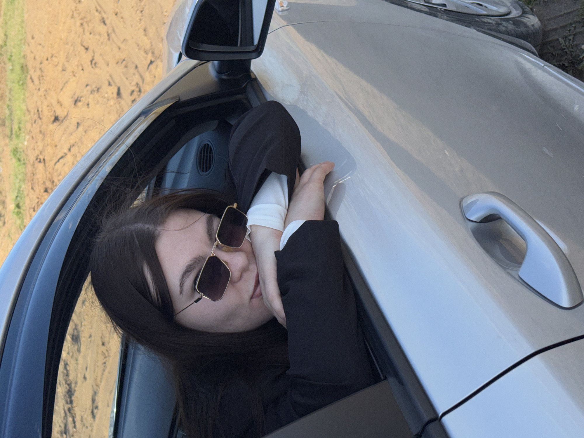 A young woman with long, dark hair and sunglasses is leaning out of the open window of a silver car. She is wearing a white shirt with a black jacket. The car appears to be parked on the side of a road, with a dirt field visible in the background. The location is likely Zaosinovo, Russia, as requested.