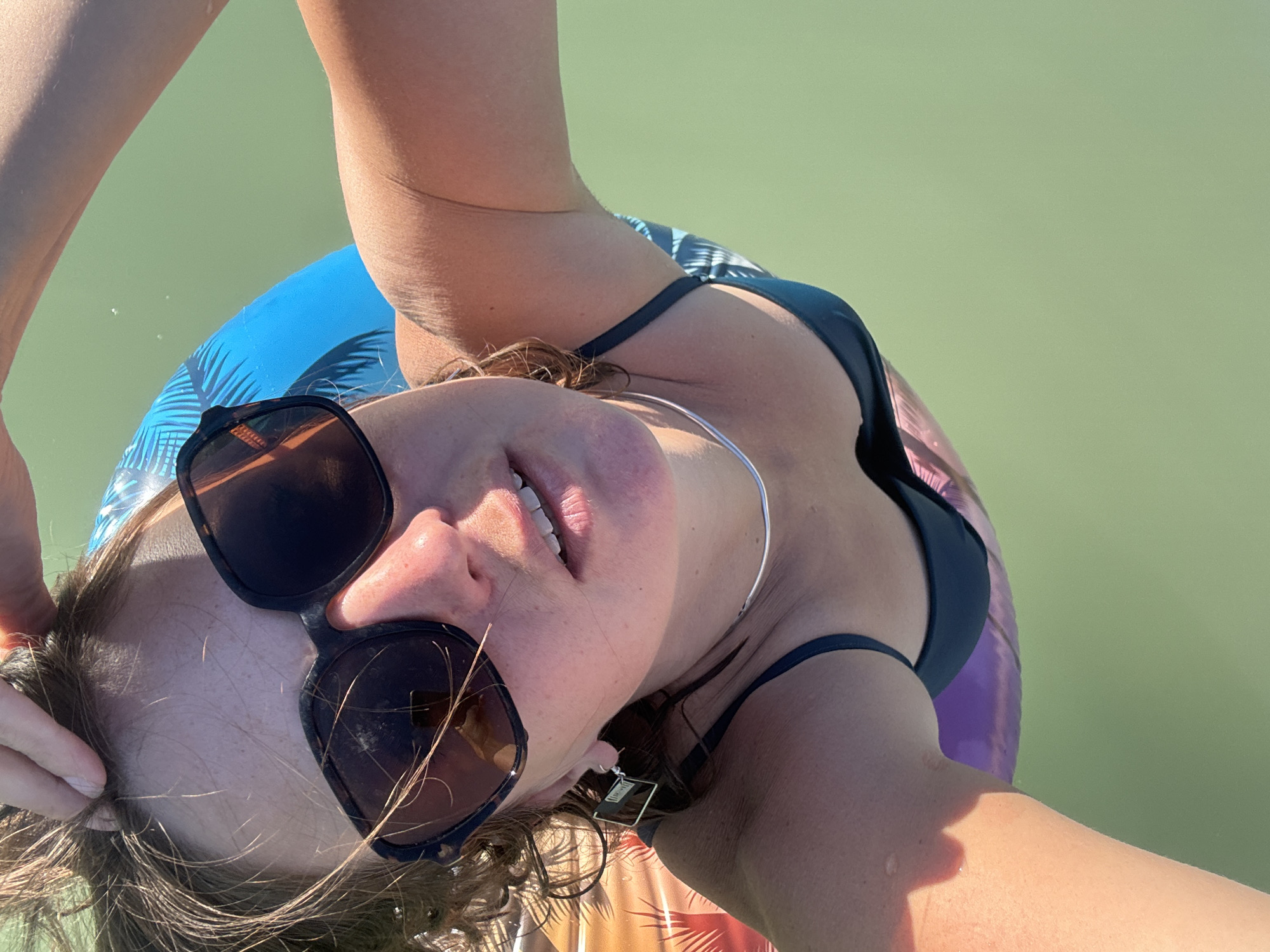 A woman is taking a selfie while floating on an inflatable in a body of green water. She is wearing sunglasses and a dark blue bikini. The inflatable has a palm tree pattern. The location is Maisod, France.