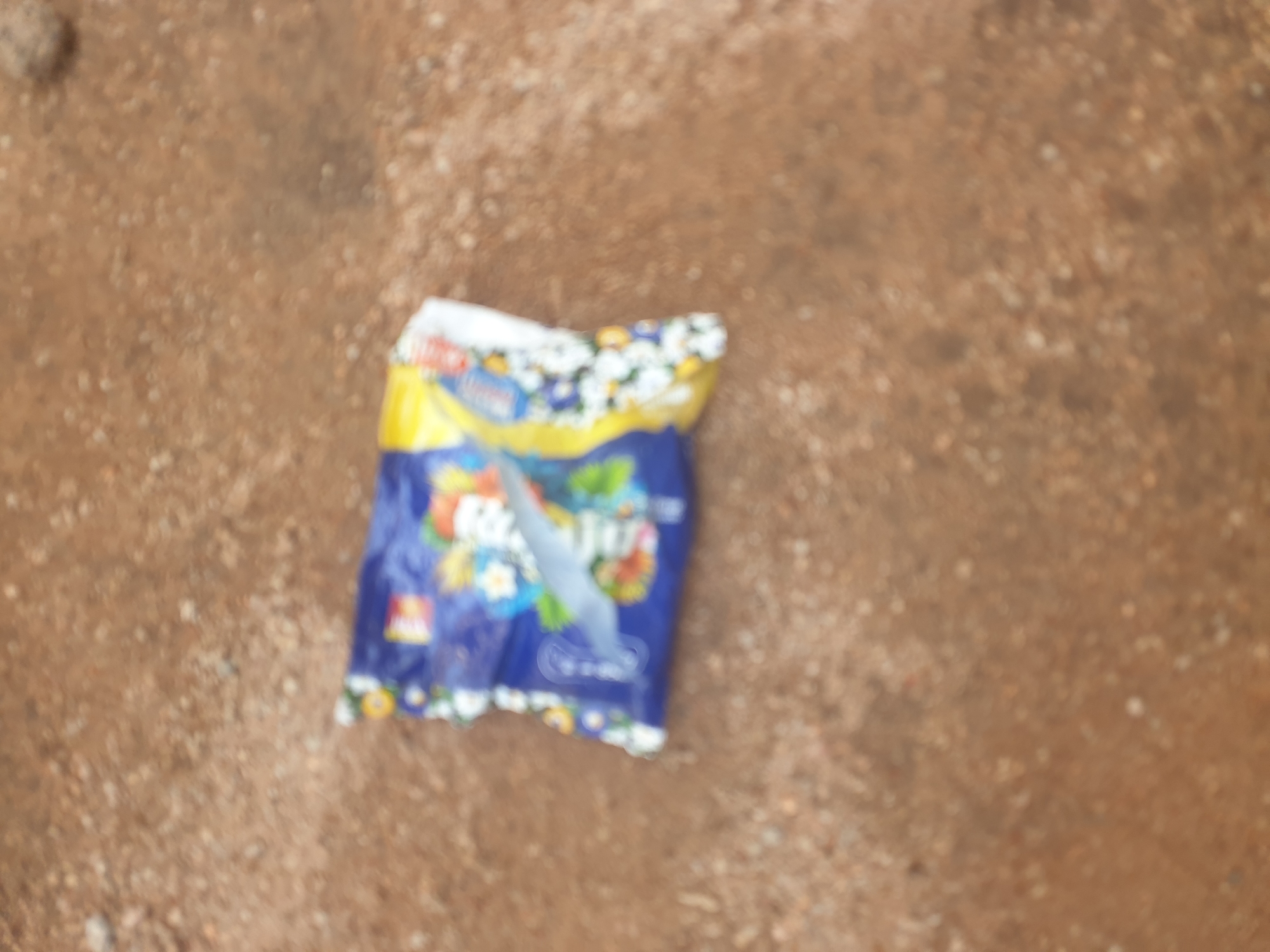 An out-of-focus, overhead shot shows a small, colorful, open bag of snacks lying on the ground. The bag is primarily blue with yellow and white accents, and appears to contain white, puffy snacks, possibly popcorn or similar. The ground is composed of small, light brown pebbles and dirt. There is no indication of human presence or any specific city or location details.
