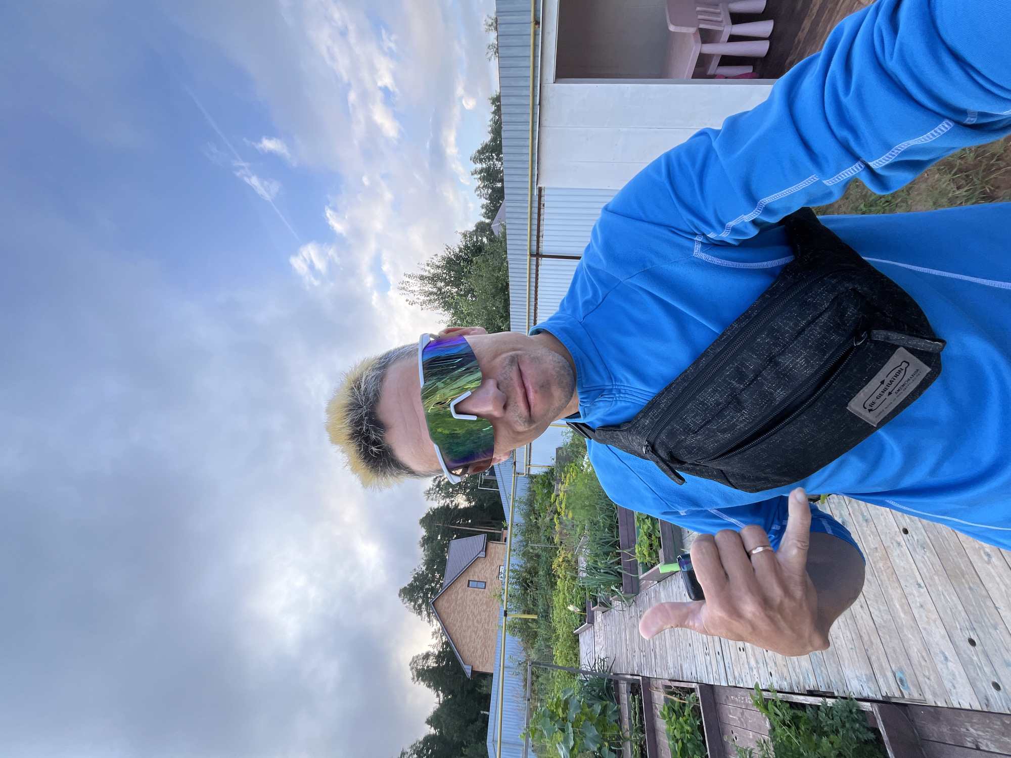 In the city of Chaikovsky, Russia, a man with blonde-tipped hair and reflective sports sunglasses poses for a selfie in a garden. He wears a bright blue long-sleeved shirt and a black fanny pack. His left hand is raised, making a "shaka" sign. The background shows raised garden beds with green plants, wooden walkways, a brick house with a gray roof, and a white corrugated metal fence. The sky is partly cloudy. A pink table and chairs are visible through an opening in the fence.