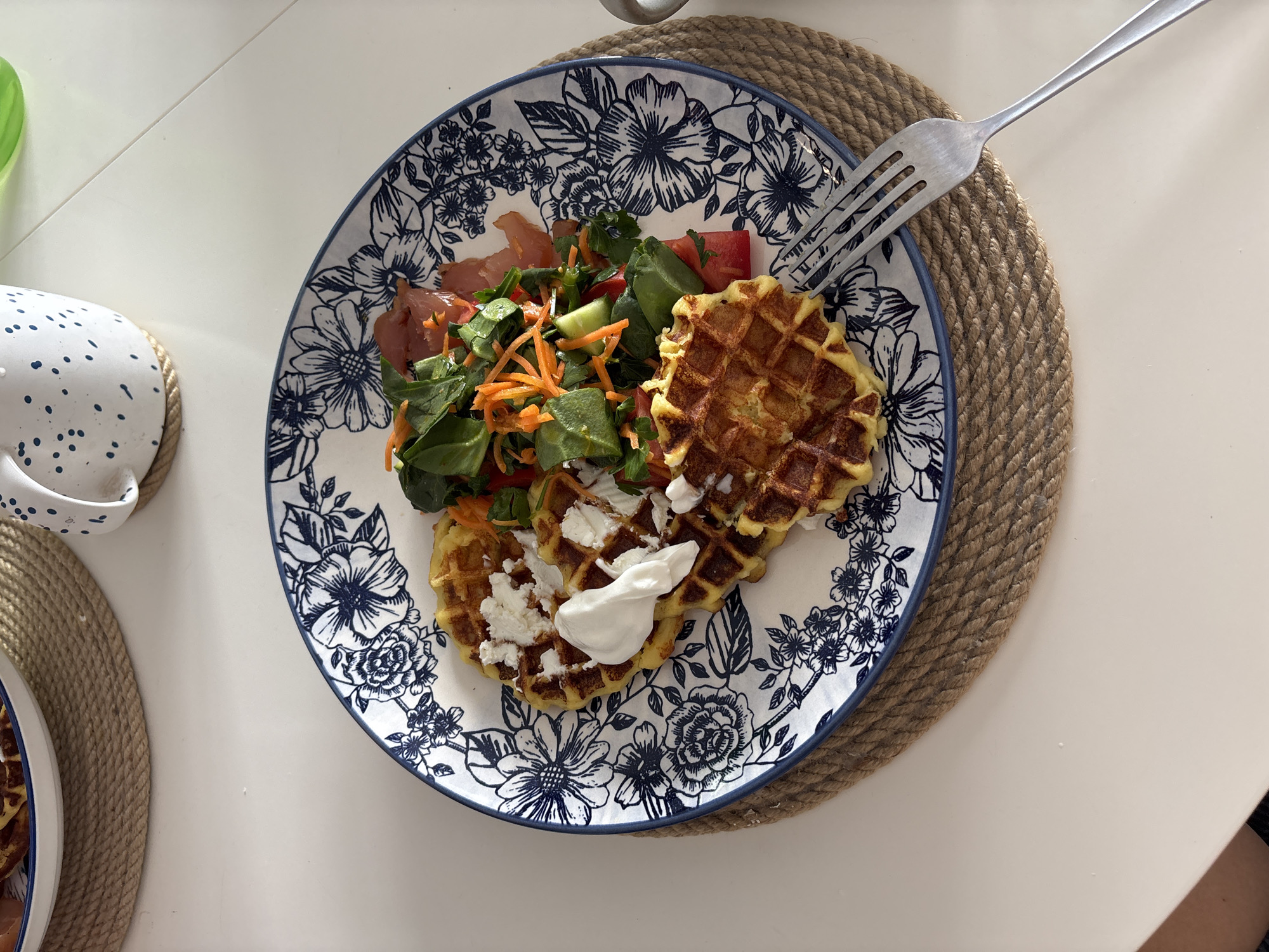 This overhead shot captures a well-composed savory meal served on a light-colored, likely white, tabletop. The central subject is a round ceramic plate, featuring an intricate blue and white floral pattern along its rim.
The plate holds three golden-brown waffles, which appear savory. Two of these waffles are topped with a generous scattering of white, crumbly cheese, resembling feta or cottage cheese. One of these also sports a dollop of smooth white cream, possibly sour cream or yogurt.
Alongside the waffles, a fresh mixed salad is arranged, consisting of vibrant green leafy greens, finely shredded orange carrots, and diced red tomatoes. Interspersed within the salad are several thin slices of reddish cured meat, consistent with prosciutto or a similar ham.
The plate rests upon a round placemat made of tightly woven, light brown natural fiber, suggesting materials like jute or rope. A silver-colored metal fork is placed with its tines resting on the upper right side of the plate and its handle extending onto the placemat.
In the upper left corner, partially visible, is a white mug or bowl adorned with blue speckles, also sitting on a similar woven placemat. The bright, even lighting indicates it is daytime. The overall scene depicts a prepared meal in an indoor setting, possibly a home dining area in Krasnoyarsk, Russia. No specific emotions or visible text are discernible.