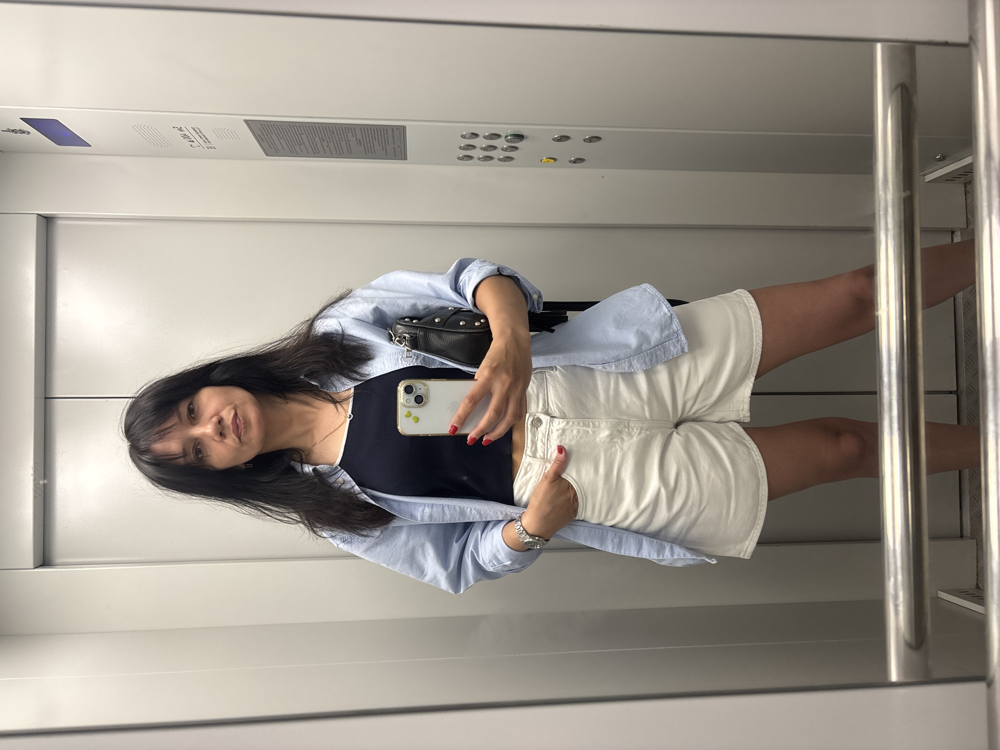A woman is standing in an elevator, taking a selfie with her cell phone. She is wearing a black tank top, white shorts, and a light blue shirt unbuttoned. A small black purse hangs from her left shoulder. The elevator has a panel of buttons, and a metal handrail is visible on the right side. The location is Unknown, Unknown.