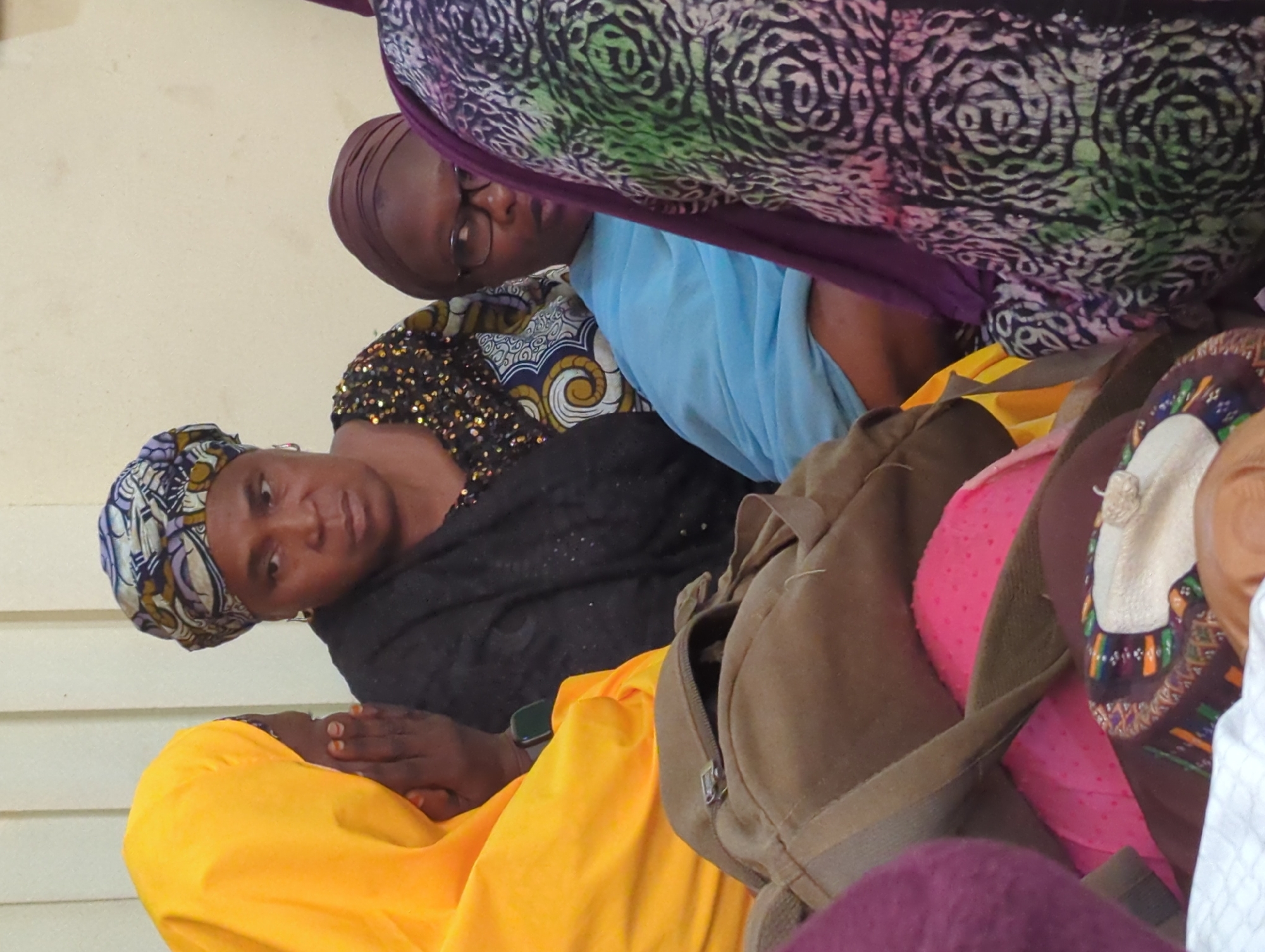 A medium shot presents several individuals indoors. A woman wearing a patterned head covering and a black shawl gazes directly at the viewer. Another woman with glasses is visible behind her, partially obscured by a patterned fabric. A person in a bright yellow garment is seated with their hands clasped. A brown backpack is positioned prominently in the lower right quadrant. The setting appears to be a crowded indoor space, although the exact location in the city of Unknown, Unknown is not discernible from the image.