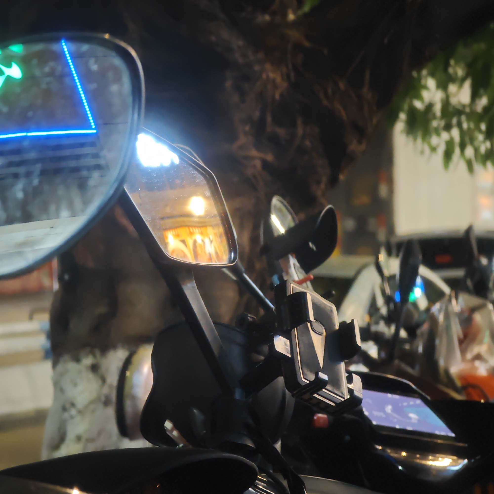 The image presents a close-up view of a scooter's handlebars and mirrors, with several other scooters visible in the background. The scene is dimly lit, appearing to be at night.
**Objects:**
* Two side mirrors of the scooter, reflecting the surrounding environment. The left mirror shows a blue neon sign, while the right shows a warm light source.
* Handlebars with a phone holder.
* Digital dashboard of the scooter.
* Other parked scooters.
* Tree and building in background.
**Environment:**
* Nighttime scene.
* Urban environment in Yuzhong District.
* Parked motorcycles, indicating street parking is available.