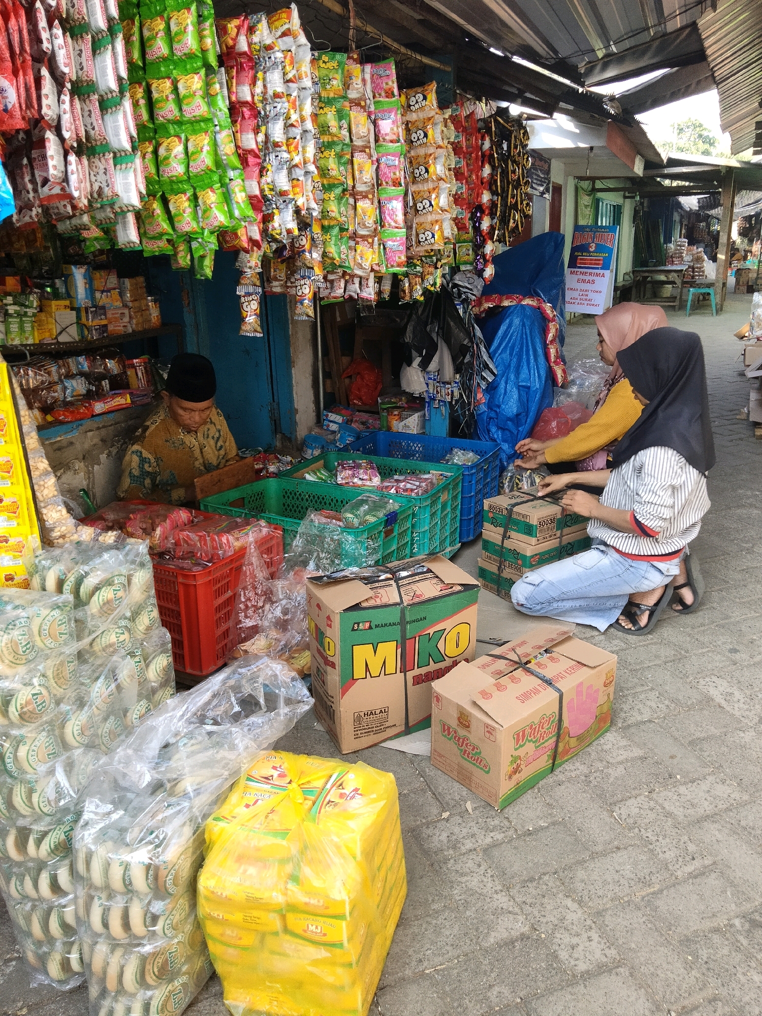 In Sawahan, Indonesia, an outdoor market stall displays an abundance of packaged snacks hanging from the roof and shelves. A person wearing a yellow shirt and headscarf is seen interacting with another person wearing a black headscarf and striped shirt, who is kneeling and tying a box with black string. Beside them, two cardboard boxes are stacked, one with green and yellow branding and the other with pink and green branding. In the foreground, large plastic-wrapped packages of snacks are piled on the ground, alongside a bright yellow plastic bag filled with more packaged snacks. A vendor, wearing a dark cap and patterned shirt, is seated behind a table laden with various goods. The scene is set on a paved ground, with the market stall covered by a corrugated roof.