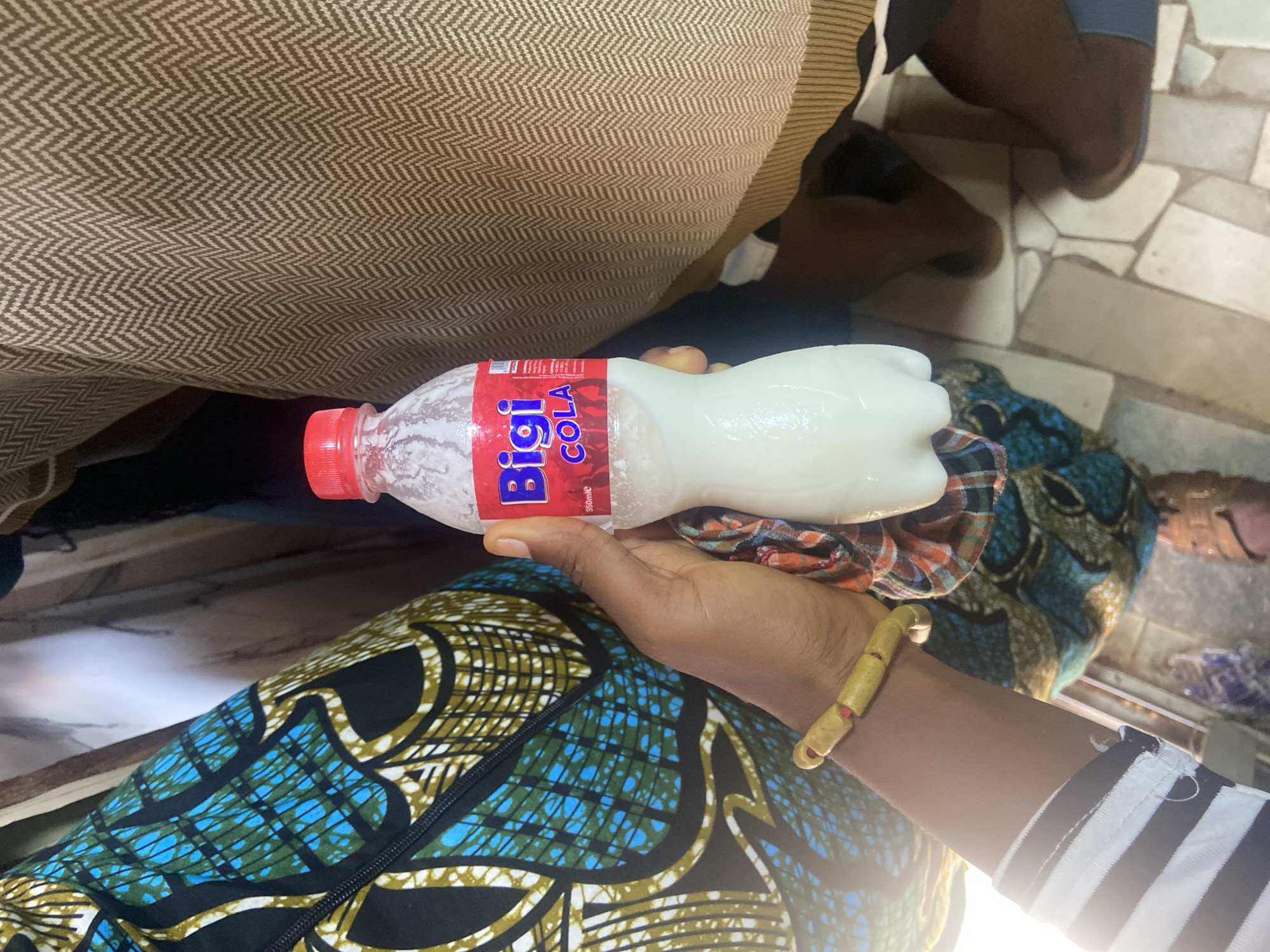 A woman is holding a bottle of Bigi Cola in Okengwe - Obehira, Nigeria. The bottle is red, white, and blue. The woman is wearing a patterned blue and black fabric over her lap. She has a yellow bracelet on her left wrist. In the background, there are other people and tiled floors. The floor is light gray. The surface under the bottle of Bigi Cola is covered in a red, blue, white and tan patterned fabric. The shirt that the woman has on is black and white striped. The Bigi Cola bottle is covered in condensation.