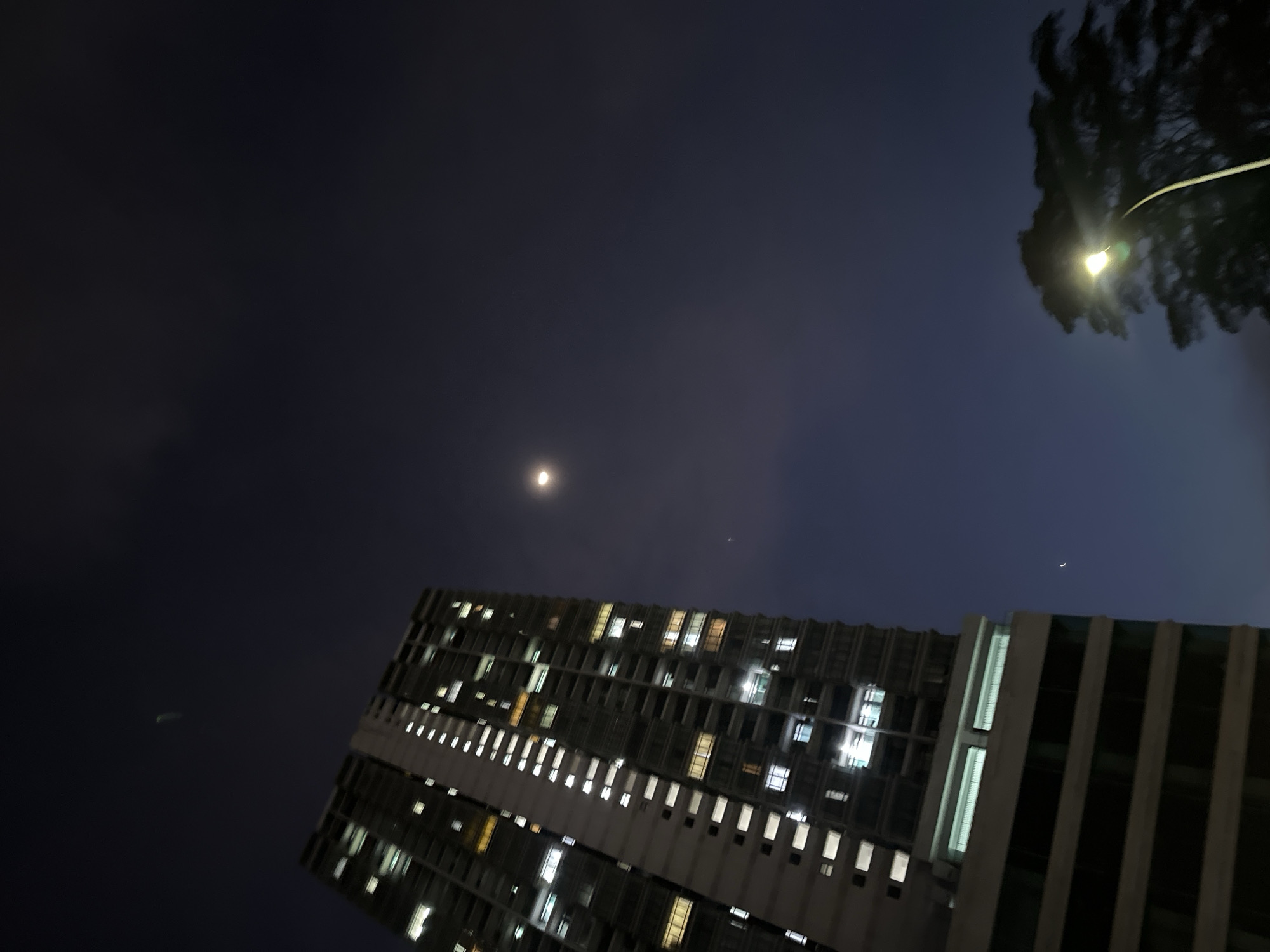 The image depicts a nighttime scene in an urban area. A tall building with multiple illuminated windows occupies the lower portion of the frame, angled upwards. Above the building, a bright, spherical object, possibly the moon, is visible in the dark sky. To the right of the frame, partially obscured by trees, a street light emits a bright glow. The sky transitions from a darker shade in the upper left to a slightly lighter hue towards the right. An unidentified aerial object is visible in the darker portion of the sky. The building's architecture suggests it might be located in a densely populated area, possibly within a city like Subang Jaya City Council, Malaysia.