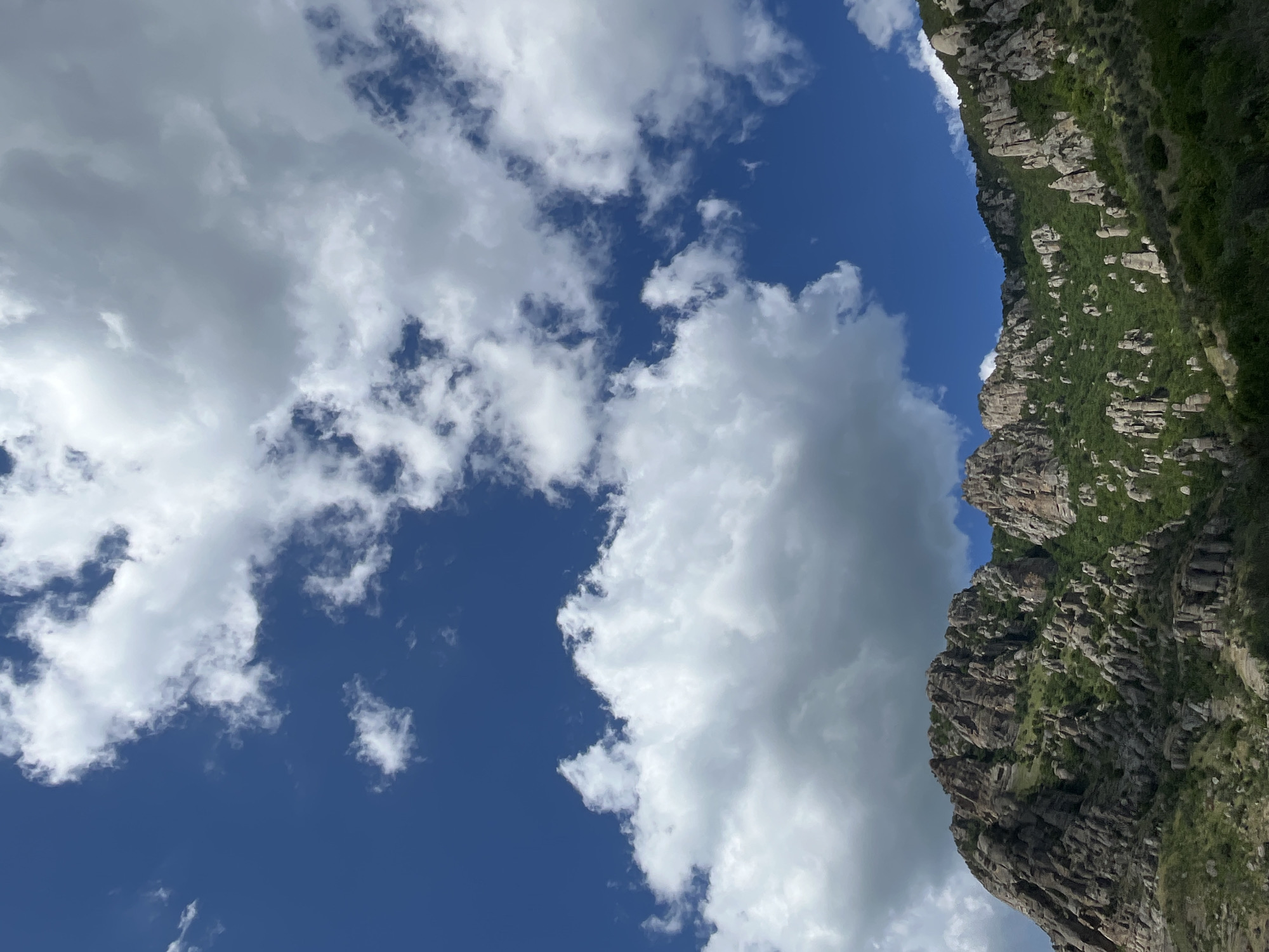 The image shows a rocky, mountainous landscape under a partly cloudy blue sky. The mountains are covered with green vegetation, and there are numerous rock formations. The clouds are white and fluffy, and they are scattered across the sky. The sun appears to be shining, as evidenced by the bright illumination of the mountains. The image is oriented vertically, with the mountains occupying the right side and the sky the left. The overall impression is one of a beautiful and rugged natural environment. The scene is serene and tranquil, evoking a sense of peace and wonder.