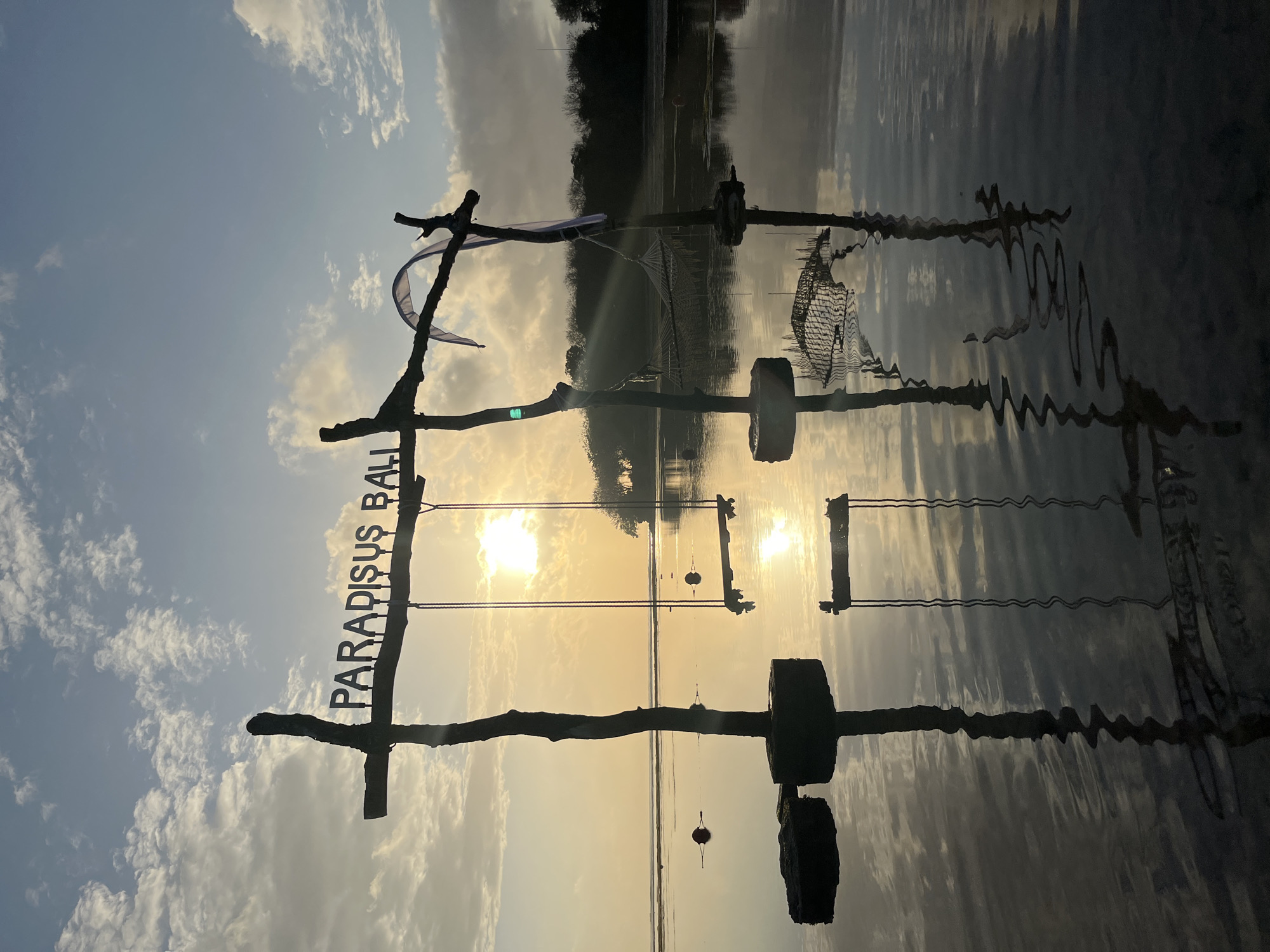 A natural light, wide-angle shot captures a sunrise over a shallow, calm body of water in Kuta Selatan, Indonesia. The sun, partially obscured by clouds, hangs low in the sky, casting a warm, golden glow. In the foreground, a wooden swing set with the word "PARADISUS BALI" emblazoned across it stands silhouetted against the vibrant sky. The swing set, made of rough-hewn branches, is anchored in the water. Reflections of the swing set, the sky, and the distant treeline shimmer on the water's surface. A hammock is visible in the background, also silhouetted. Several buoys are scattered across the water. The overall scene evokes a serene and peaceful atmosphere, characteristic of a tropical sunrise.