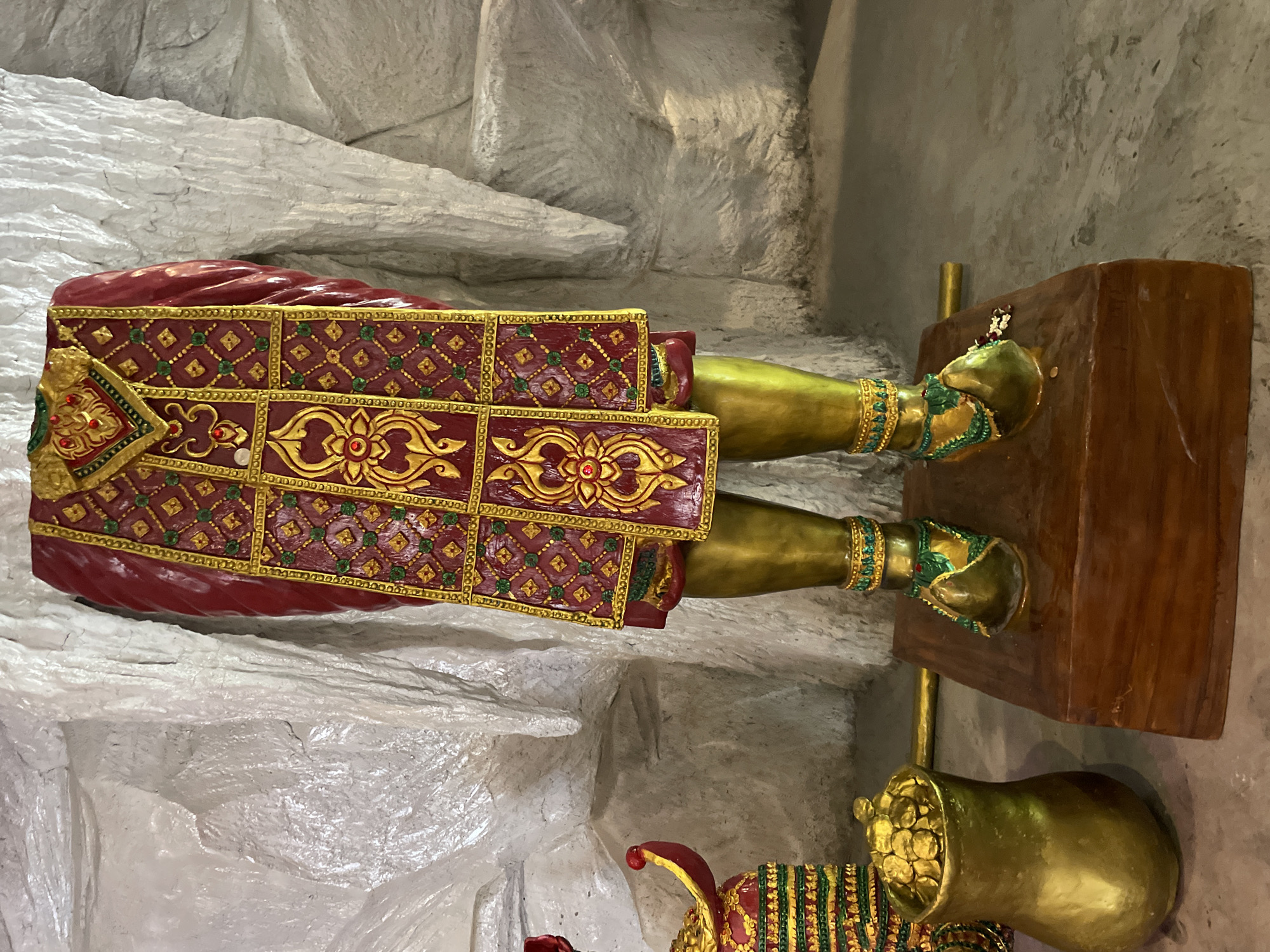 The image shows a statue of a figure, possibly a deity or royalty, from the waist down. The statue's legs are golden, and it wears ornate, jeweled anklets and sandals. The lower torso is adorned with a richly decorated skirt or robe in deep red with intricate gold and green patterns, featuring floral motifs and geometric designs. The background appears to be a rough, textured, light-colored wall, possibly made of plaster or stone. To the left of the statue, a golden pot filled with what appears to be gold coins is visible. The statue is standing on a wooden platform. The overall impression is one of opulence and religious or cultural significance. The image was taken in Bo Win Subdistrict, Thailand.