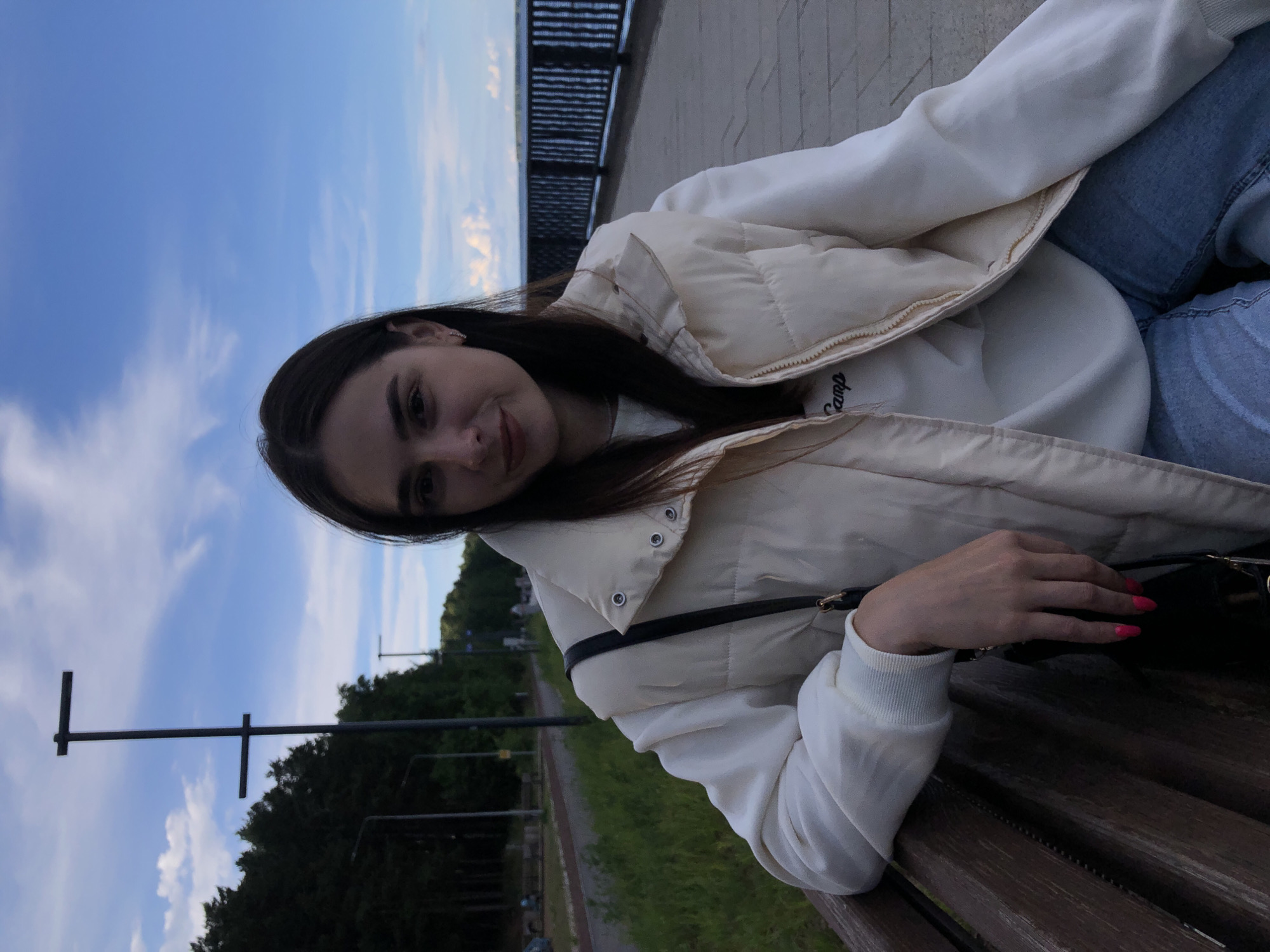 A young woman is sitting on a bench in Chaikovsky, Russia. She is wearing a white vest, a white sweatshirt, and blue jeans. She is looking at the camera and smiling slightly. There is a black bag sitting on the bench next to her. In the background, there is a body of water with a black fence, a park, and a blue sky.