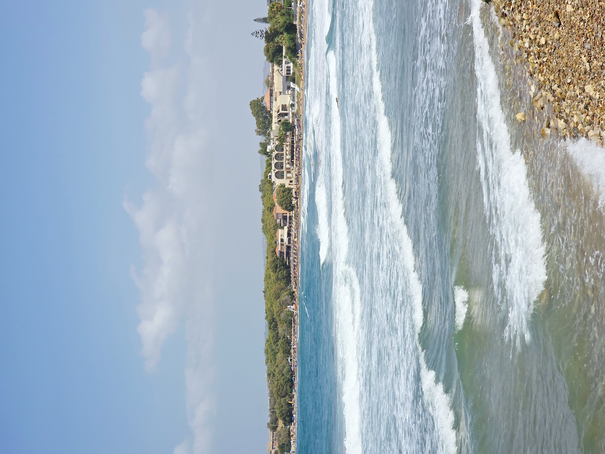 The image depicts a scenic view of a beach and the sea in Daratsos, Greece. The foreground features a pebbly shoreline with a gentle wave lapping at the edge. In the mid-ground, the turquoise sea is filled with white-capped waves rolling towards the shore. In the background, a crescent-shaped beach is lined with numerous umbrellas and sunbathers. Behind the beach, lush green trees form a dense backdrop, with a few buildings, possibly hotels or resorts, visible nestled amongst the foliage. The sky above is a clear, bright blue with a few scattered white clouds. The overall impression is one of a tranquil and picturesque coastal resort.