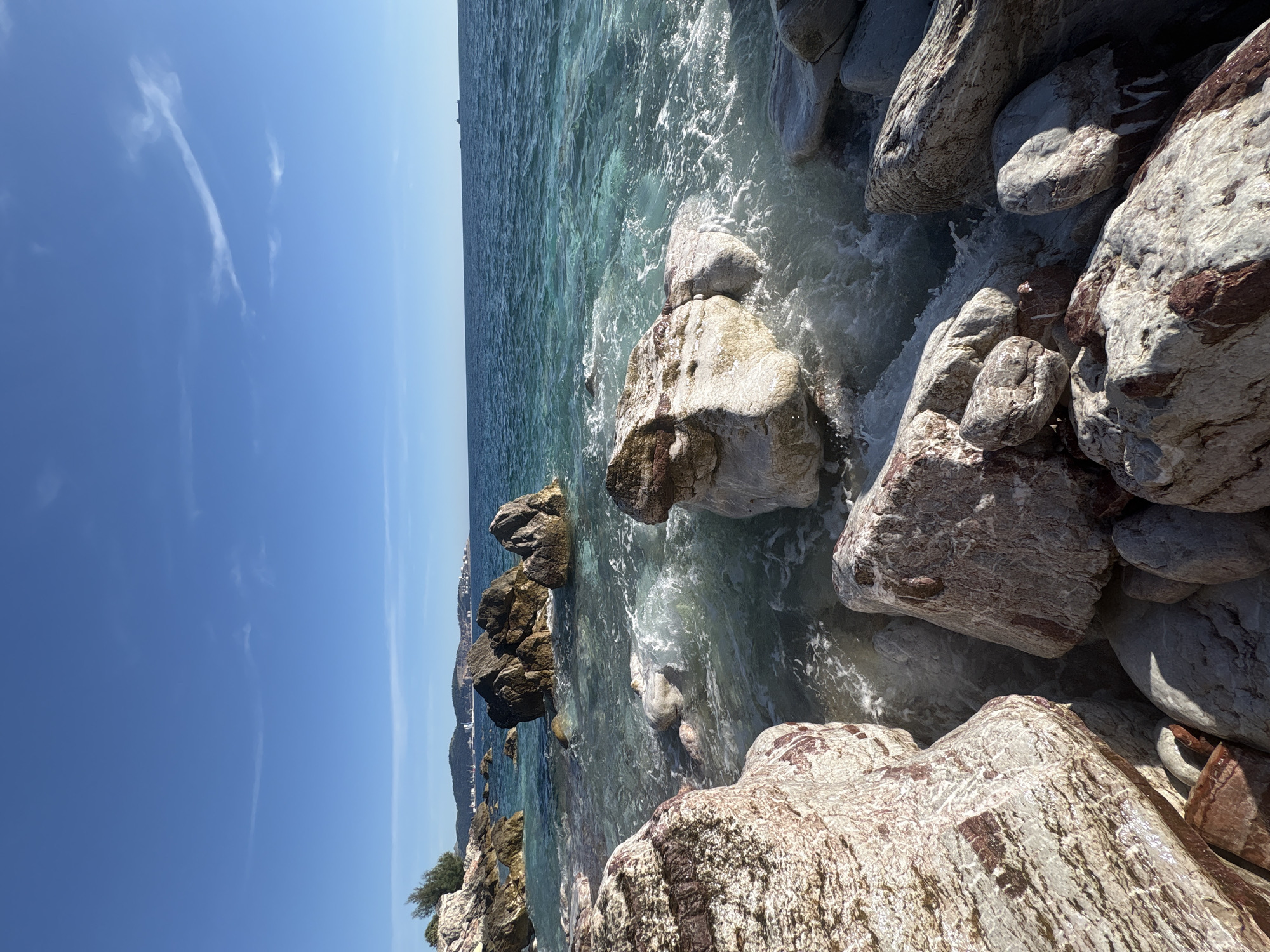 The image captures a rocky coastline along the Adriatic Sea in Bar, Montenegro, under a clear, bright blue sky. The water is turquoise and translucent, with waves gently lapping against the large, weathered rocks. The rocks are light grey and reddish-brown, displaying a rugged texture. In the distance, the coastline of Bar is visible, with buildings and hills visible along the coastline. The scene is peaceful and serene, portraying a typical Mediterranean landscape.