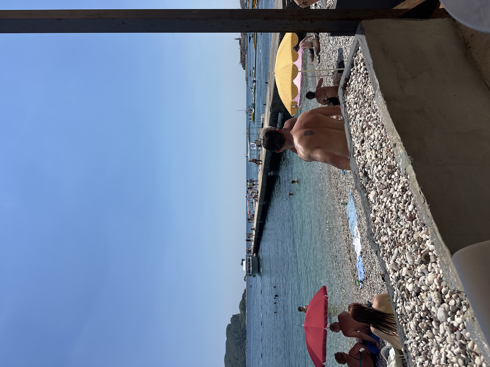 A sunny day at a pebbled beach in Budva, Montenegro, features several people enjoying the water and the shore. In the foreground, a textured concrete planter filled with small stones is visible. To the left, a red umbrella shades a seated couple. Further out, people swim in the clear blue water, and a long pier extends into the sea, populated by individuals and a white boat docked at its end. In the distance, a town with red-tiled roofs sits on a peninsula, and a rocky island is visible to the far left. The sky is clear and blue.
