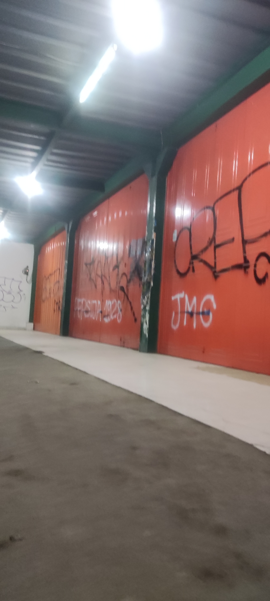An outdoor scene shows an orange corrugated metal wall with graffiti. The wall is divided into sections by vertical dark green beams. The graffiti on the wall includes the words "PERSIDA 1928" in white, and "JMG" also in white. To the left, there is another section of wall that is white and has some black graffiti. A section of the roof is visible at the top, supported by dark beams. Fluorescent lights are mounted on the roof, illuminating the scene. The ground in the foreground is a mix of gray asphalt and a lighter colored paved area. This scene is located in the city of Gebang, Indonesia.