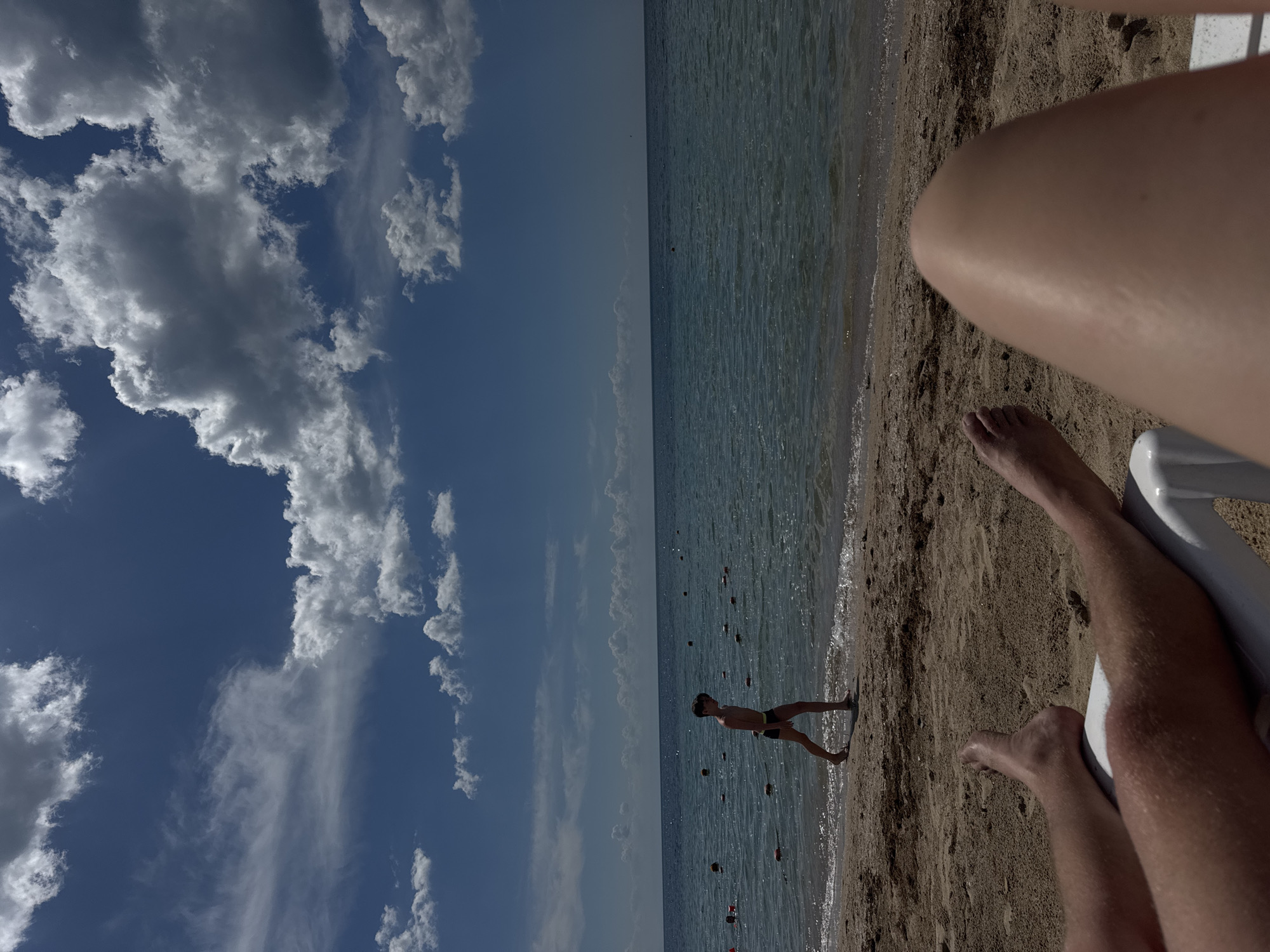 A horizontal-orientation photograph captures a coastal scene, presenting a composition of sky, sea, and beach. In the distance, a child is walking along the shoreline. Nearby, a fair-skinned adult reclines in a white beach chair. The sky is a clear blue with scattered white clouds. The water is blue, with small waves visible along the shore. Red buoys can be seen along the shoreline. The photograph suggests a beach setting, possibly in Yevpatoriya, Ukraine.
