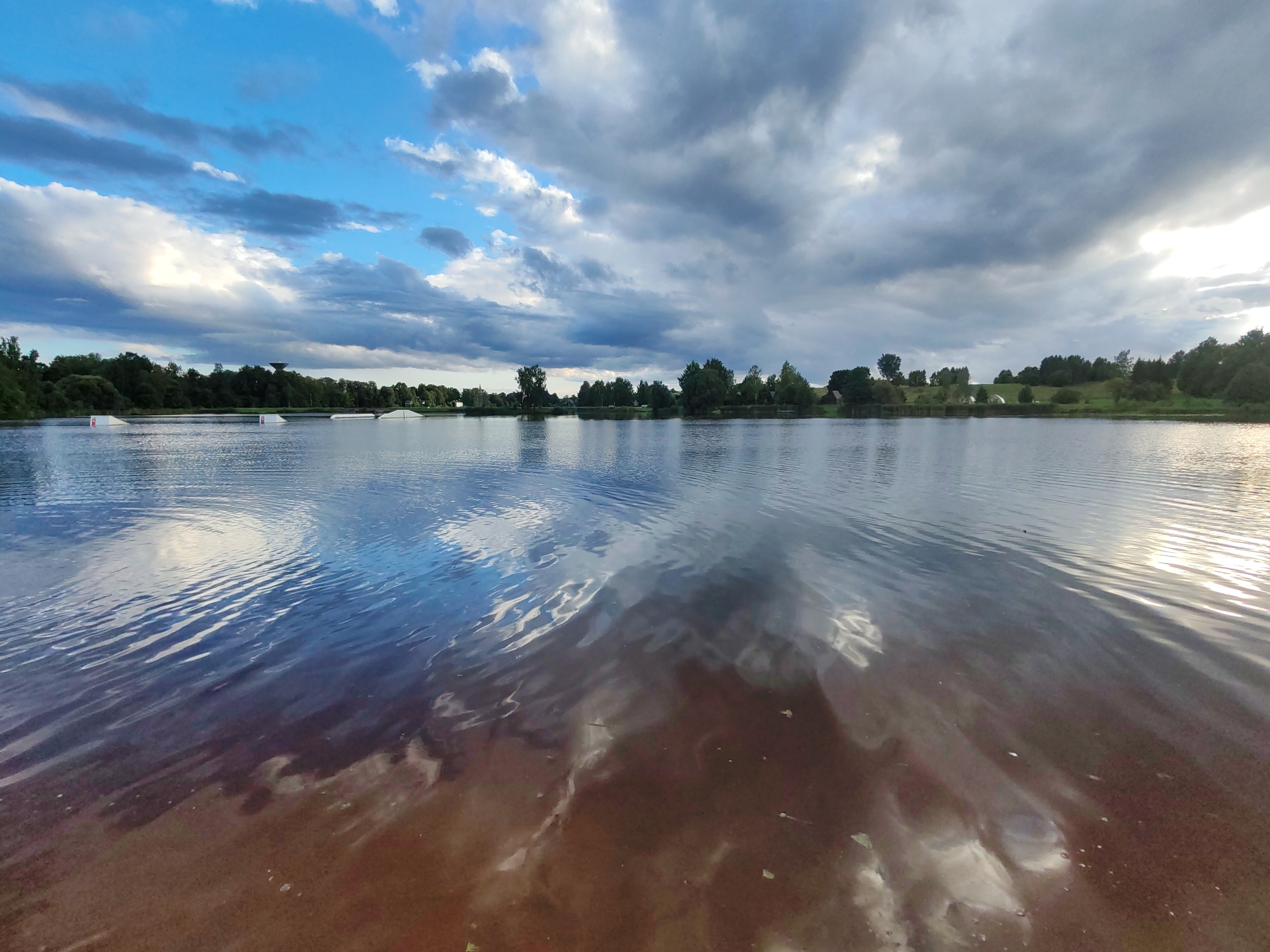 The image shows a wide-angle, eye-level view of a lake with a tree-lined shore. The sky is filled with dramatic clouds, with patches of blue sky visible. The water reflects the sky and surrounding trees. On the left side of the lake, there are several white structures that appear to be part of a water sports facility, possibly for wakeboarding or similar activities. The shore is covered in green grass and trees, with some buildings visible in the distance. The overall impression is of a natural landscape within or near a city, likely during the late afternoon or early evening due to the lighting.